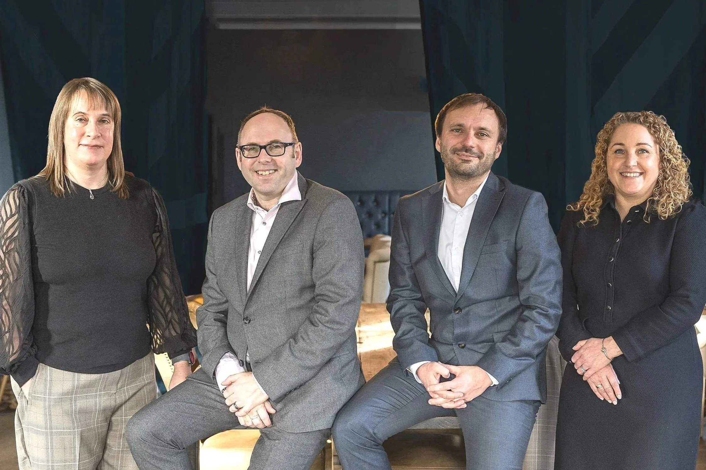 Four professionally dressed people, two men and two women, looking into the camera.  The two men in the middle are perched on the end of a table and the two women are standing either side of them.