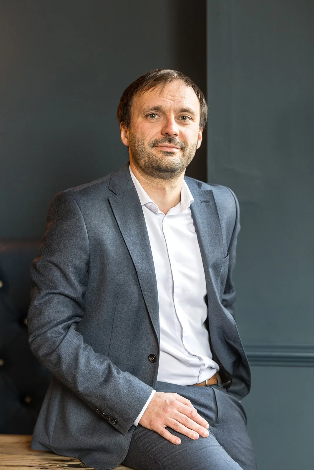 A professionally dressed man with brown hair and a beard.  He's wearing a suit and is leaning on the side of a table