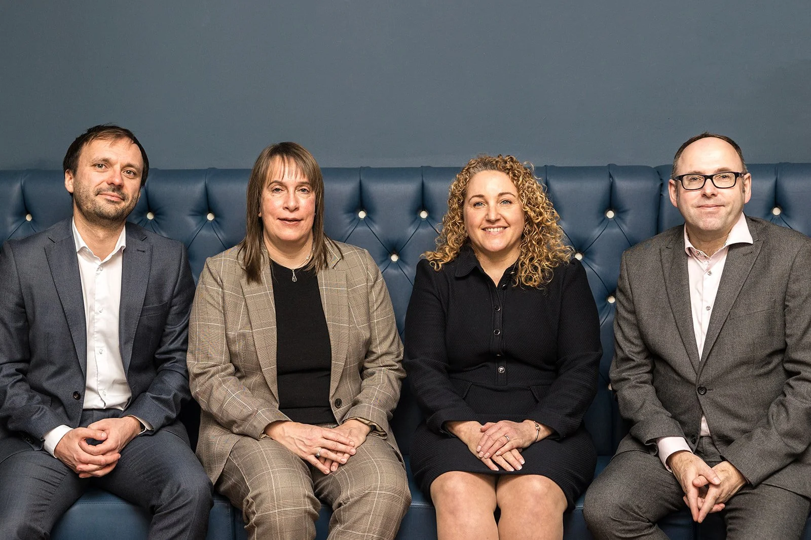 Four people, two women and two men, sat side by side on a padded bench.  They are professionally dressed.