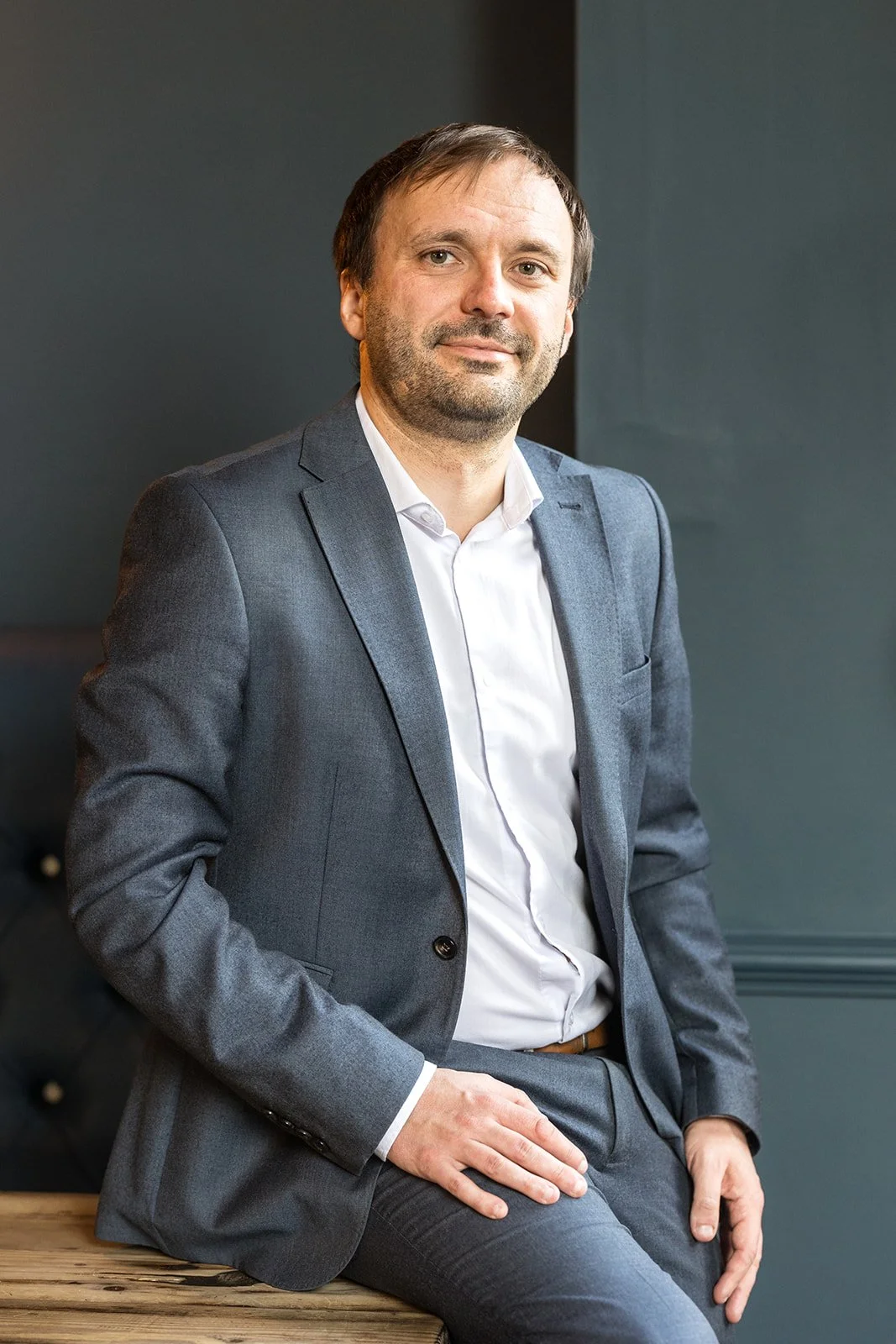 A man wearing a gray suit and white shirt, sitting on a wooden surface with a dark wall behind him.
