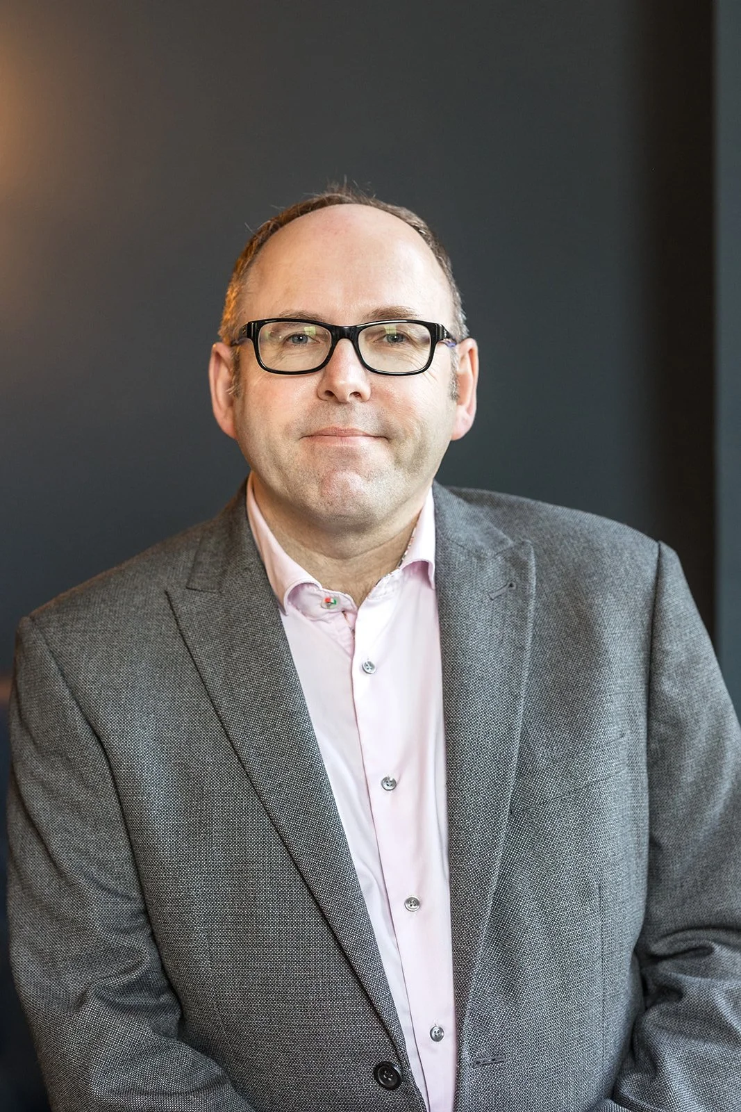 A professional portrait of a middle-aged man with glasses, wearing a gray blazer and a light pink shirt, standing against a dark background.