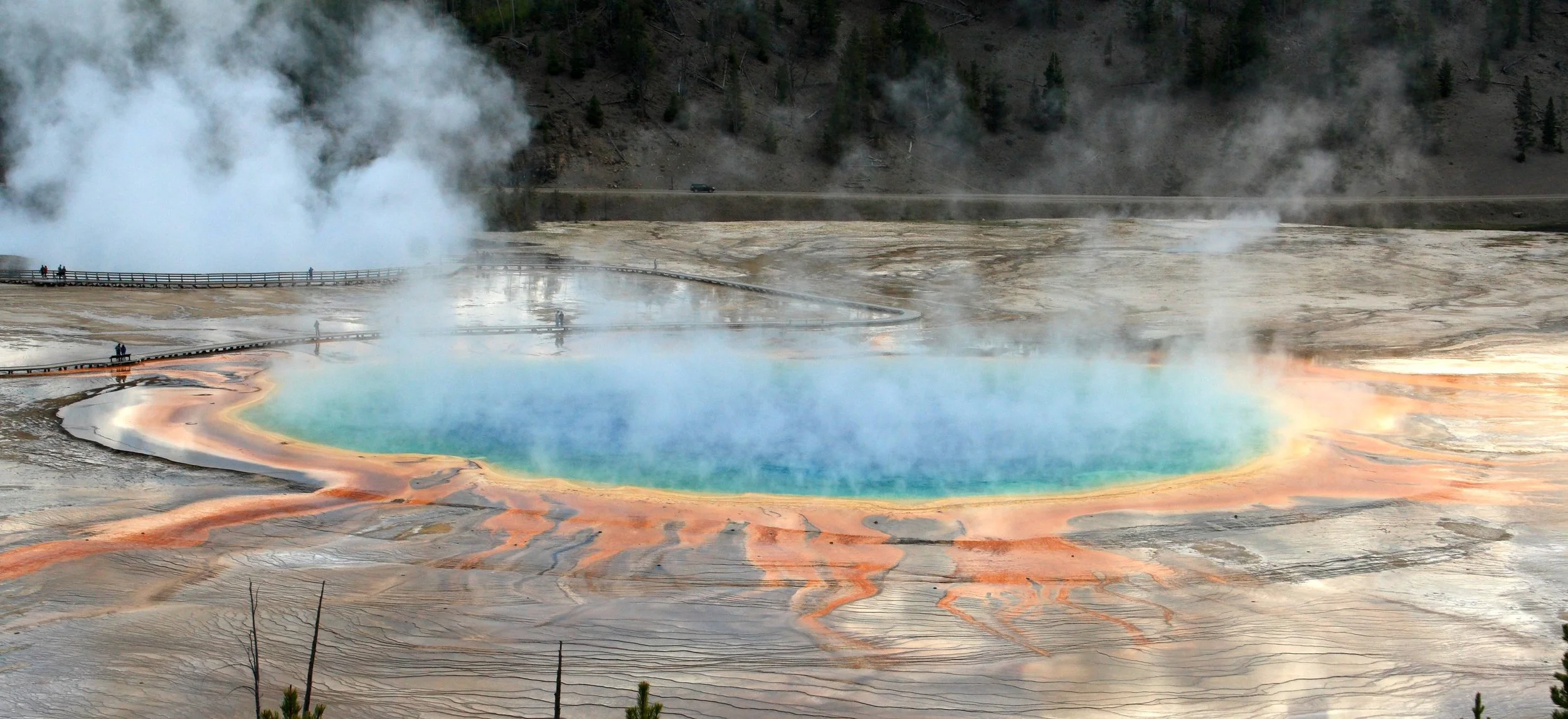 Colorful hot spring with steaming water, surrounded by mineral deposits in yellow, orange, and brown hues, and a boardwalk with people walking around.
