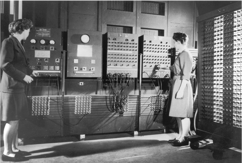 Two women working with vintage early computer or calculator machinery, surrounded by large panel boards and switches, in an industrial or office setting from the mid-20th century.