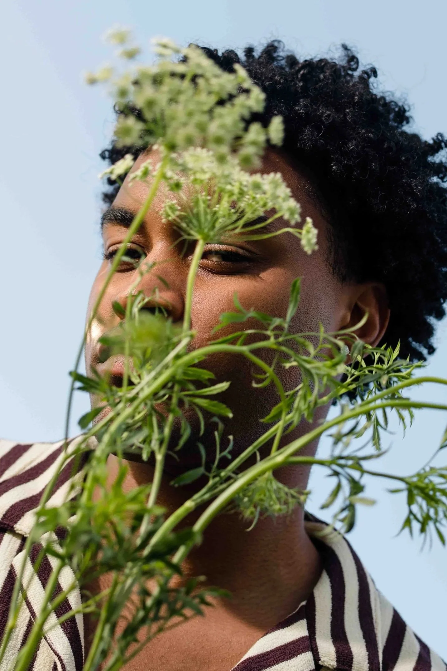 A person with short curly hair holding a large green plant with white flowers in front of their face, smiling slightly against a blue sky background.