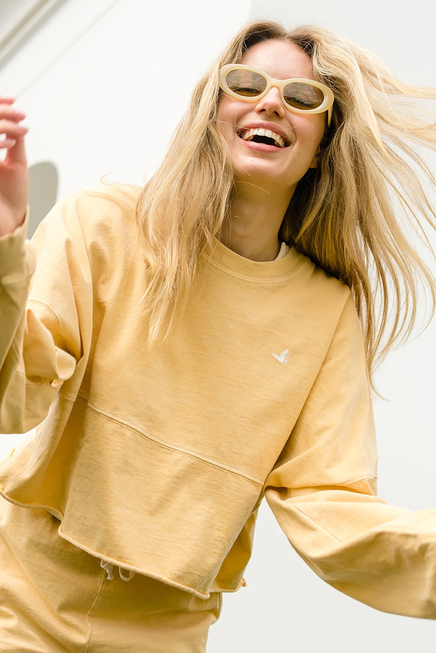 A woman with long blonde hair wearing yellow sunglasses, a yellow sweatshirt, and matching pants, smiling and enjoying herself outdoors.