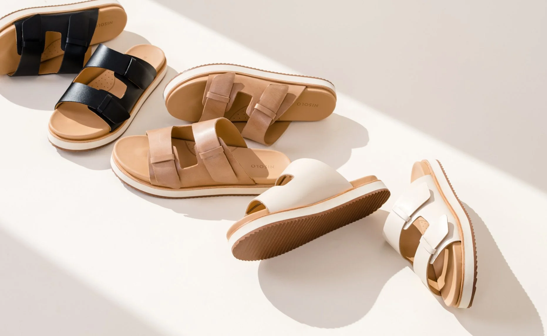 A collection of six pairs of slide sandals in black, beige, and white colors arranged on a white surface with sunlight casting shadows.