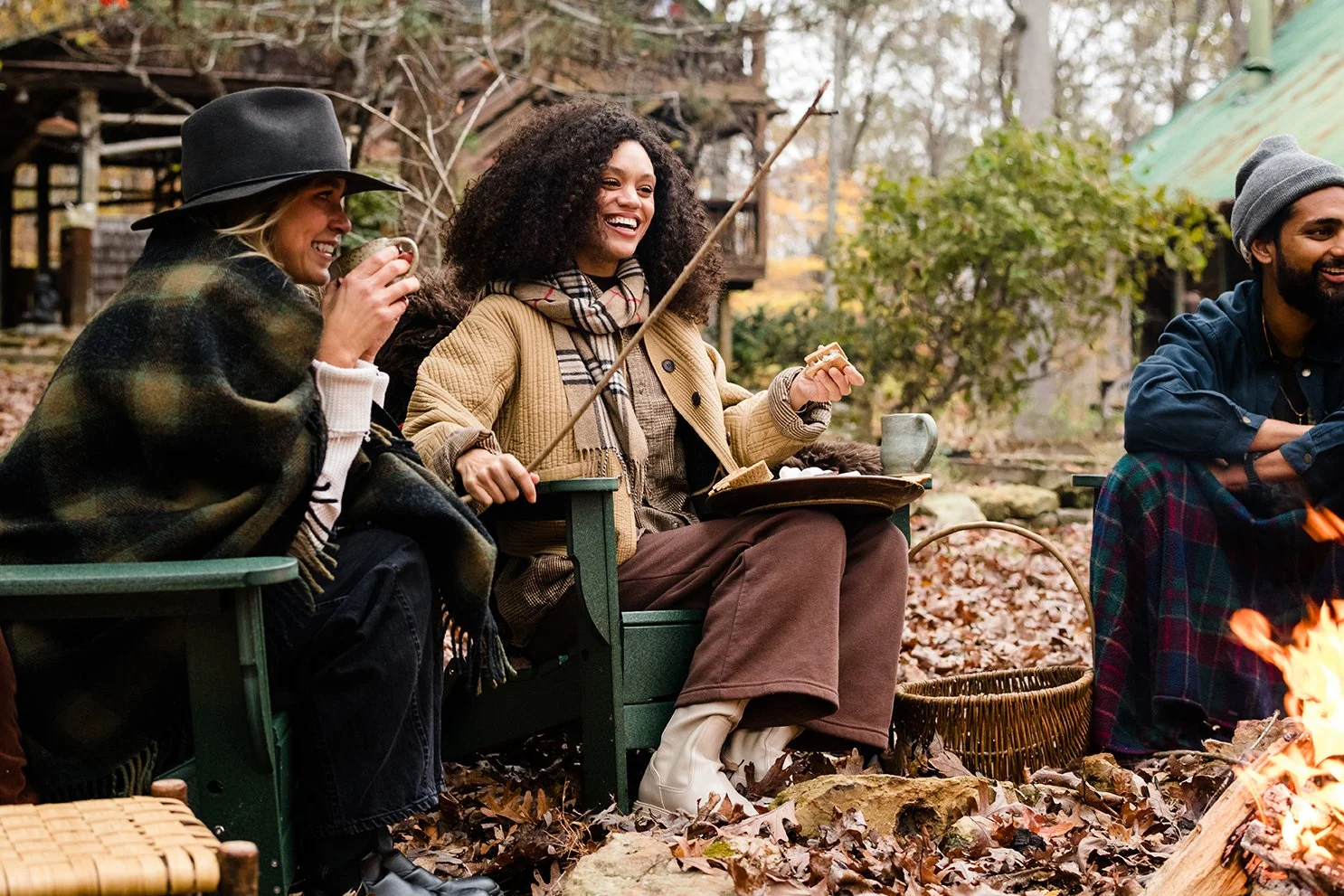 Three friends sitting outdoors near a campfire, enjoying snacks and hot drinks on a fall day.