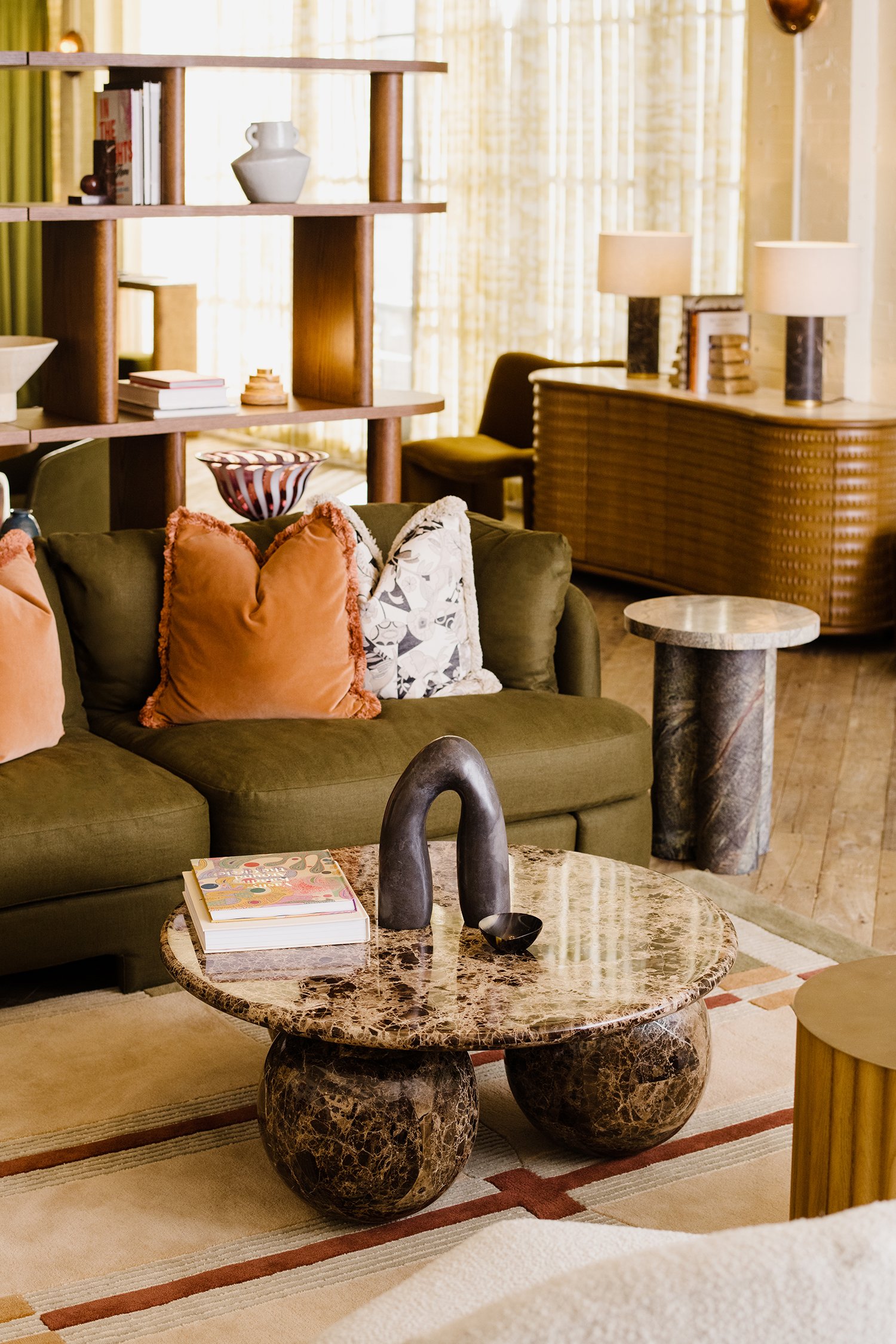 Living room with green sofa, orange and patterned pillows, marble coffee table with sculpture and books, wooden shelves and sideboard, lamps, and window with curtains.