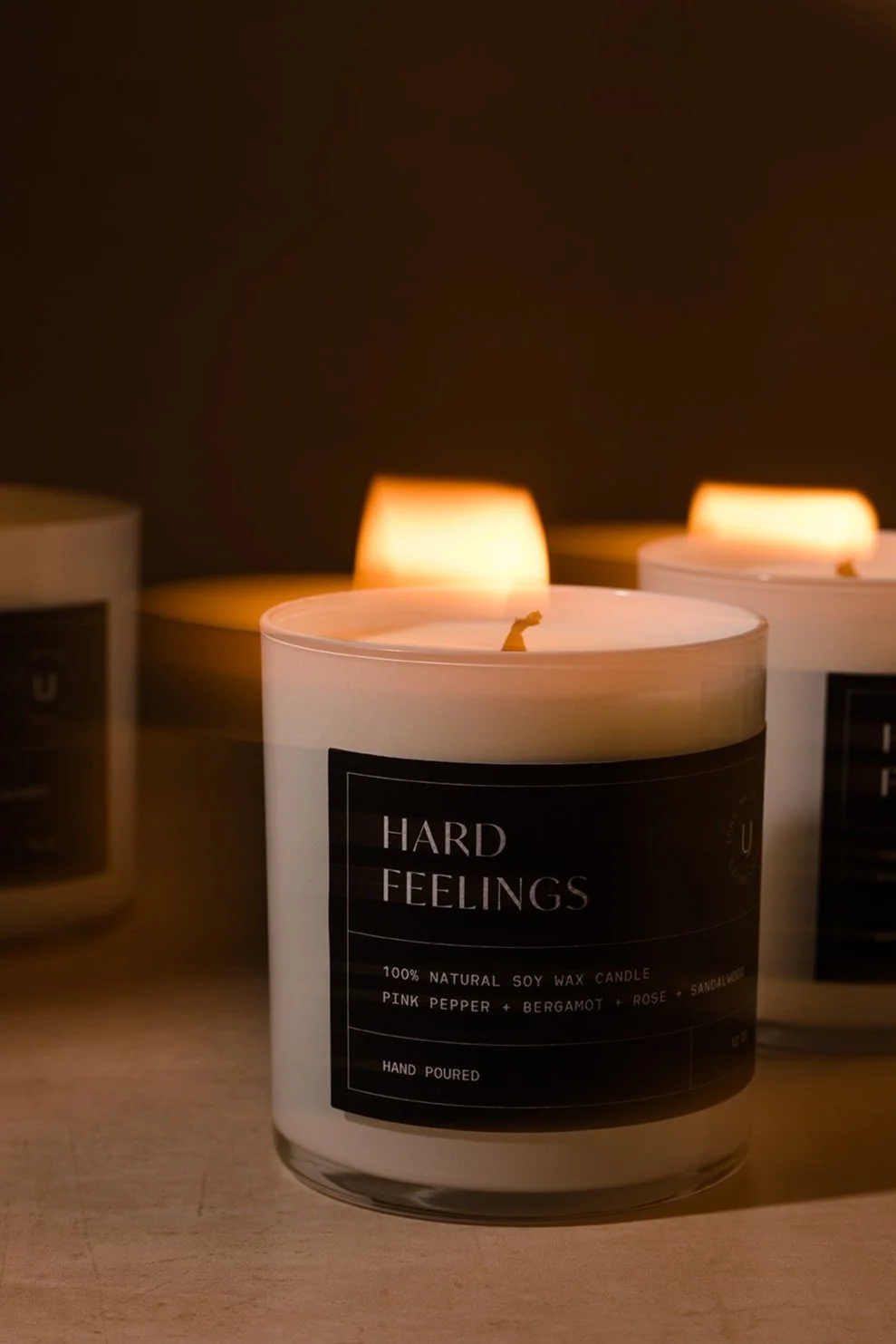 A lit candle labeled 'Hard Feelings' with a scent of pink pepper, bergamot, rose, and sandalwood, made of natural soy wax.