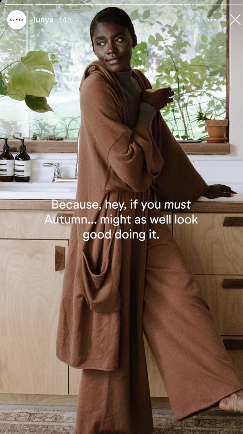 A woman in a brown outfit leaning against a wooden kitchen counter with a large window, green plants, and bottles on the counter behind her.