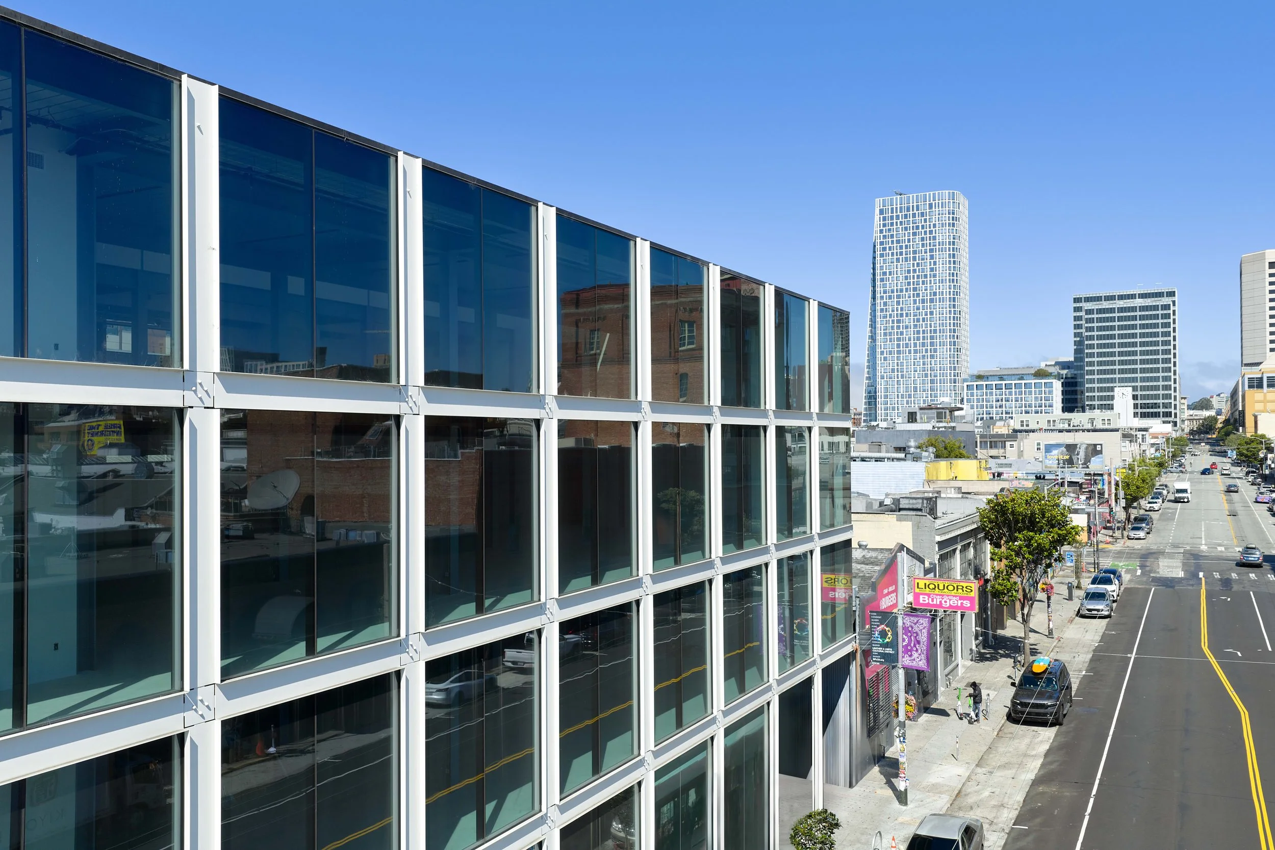 City street view featuring a modern glass building on the left, a clear blue sky, and various high-rise buildings in the background. The street has parked cars, sidewalks with trees, and storefronts including a liquor store and a burger restaurant.