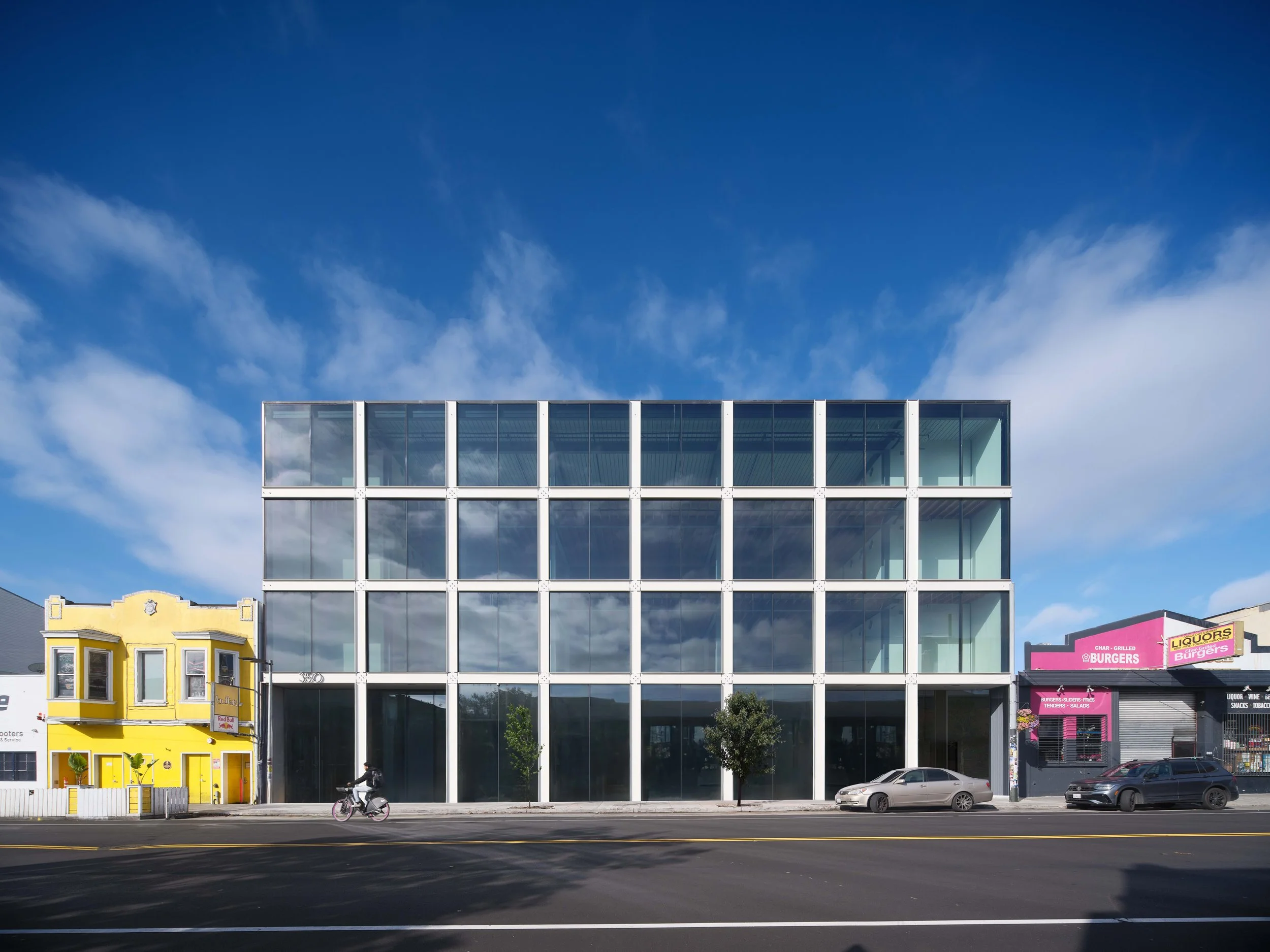 Modern glass building with a blue sky and clouds behind it, yellow building on the left, and a black building on the right with a pink sign, cars parked in front, and a person riding a bike on the street.