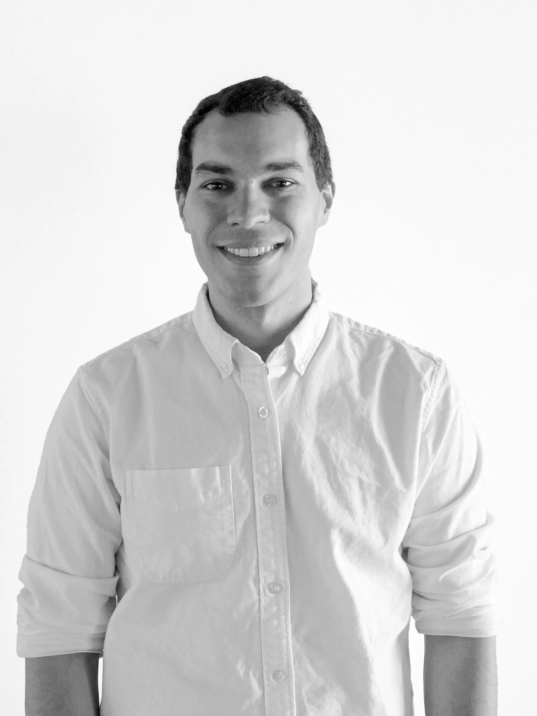 Black and white photo of a smiling young man wearing a button-up shirt against a plain white wall.
