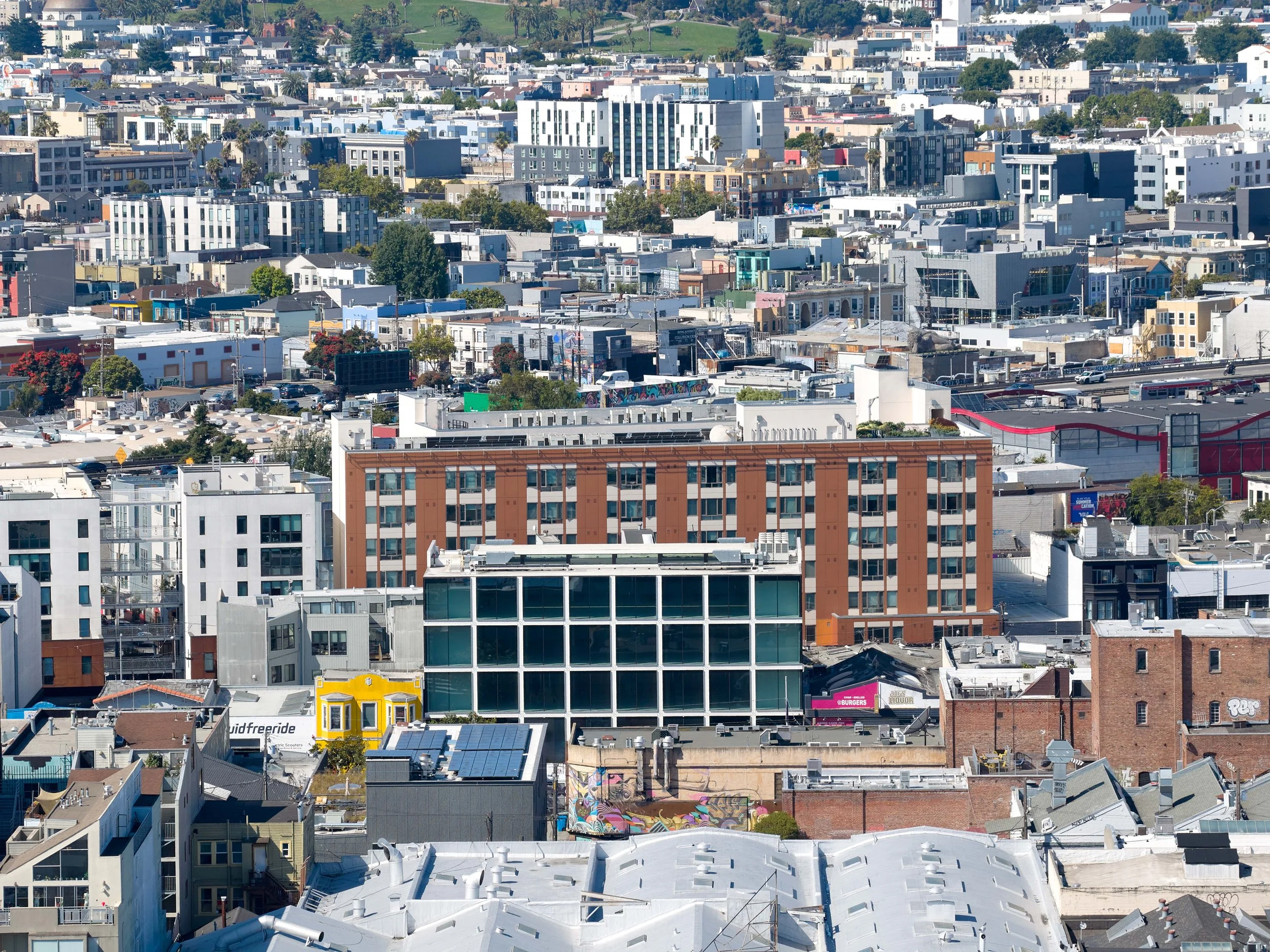 Aerial view of a dense cityscape with various modern mid-rise and high-rise buildings in different architectural styles and colors, under a clear blue sky.