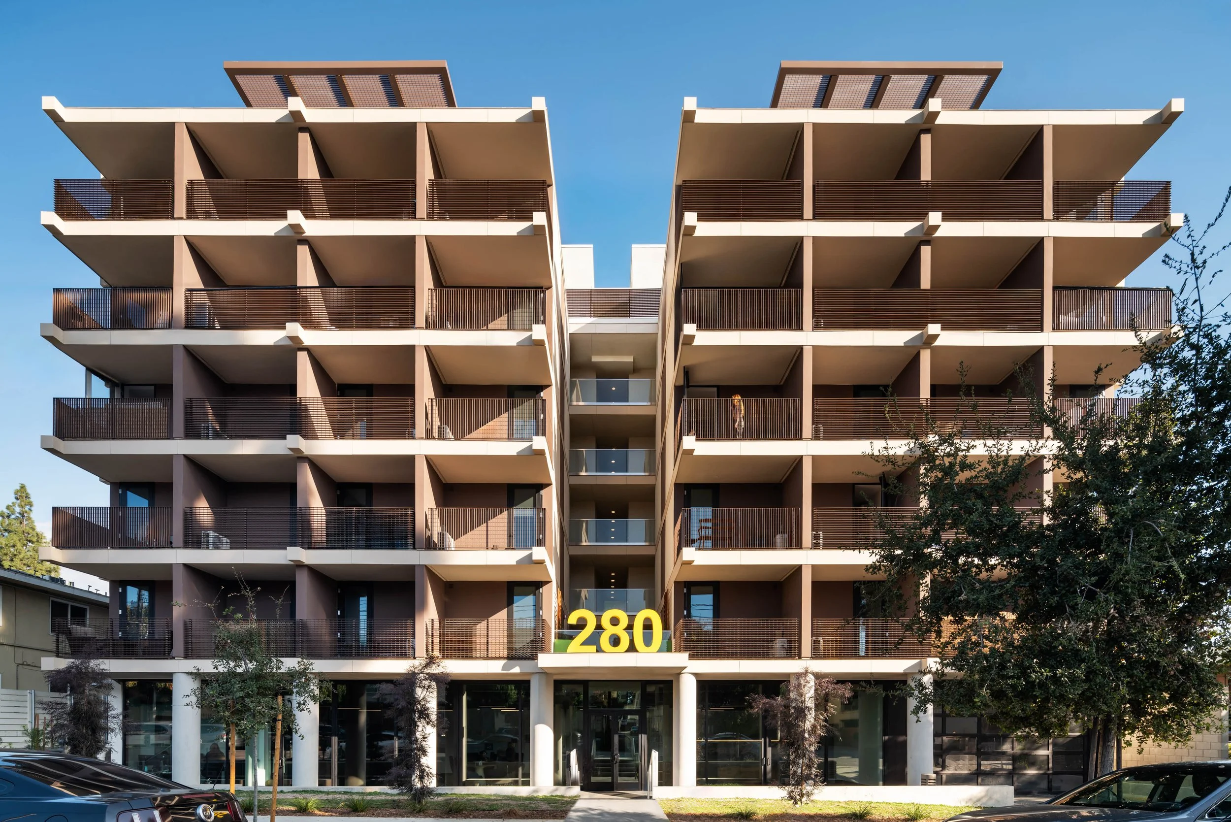 Modern multi-story apartment building with seven floors, featuring balconies with brown railings, a glass entrance, and the number 280 in large yellow digits above the entrance. Trees and parked cars are visible in front of the building.