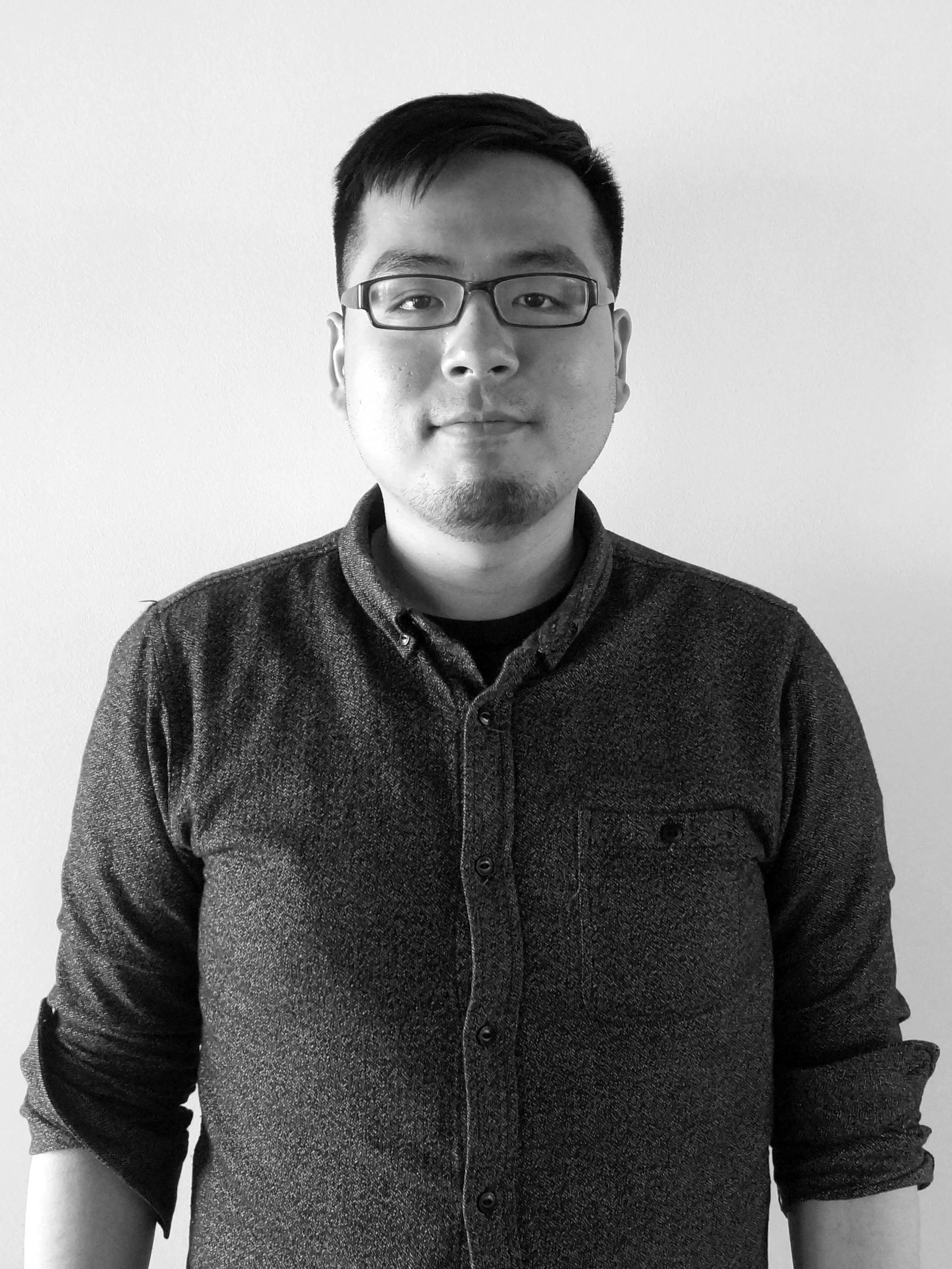 Black and white portrait of a young man with glasses and short dark hair, wearing a collared shirt with rolled-up sleeves, standing against a plain wall.