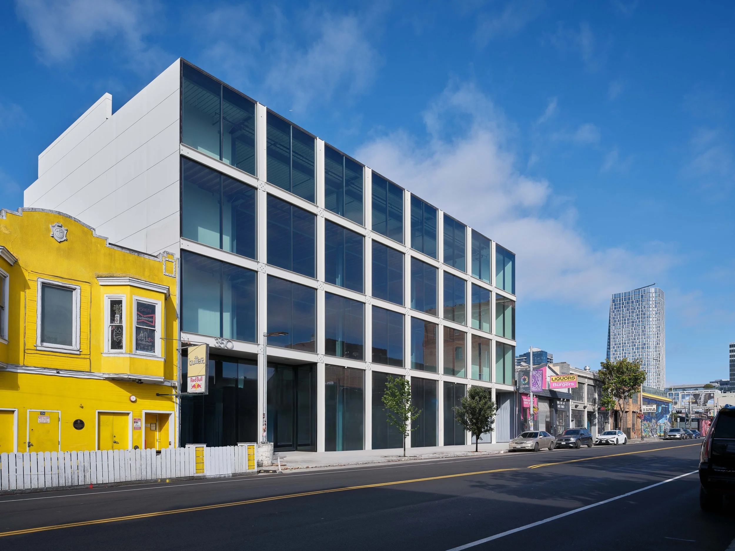 A modern glass-front building beside a yellow old-style building on a city street with few cars and small trees, under a blue sky with some clouds.