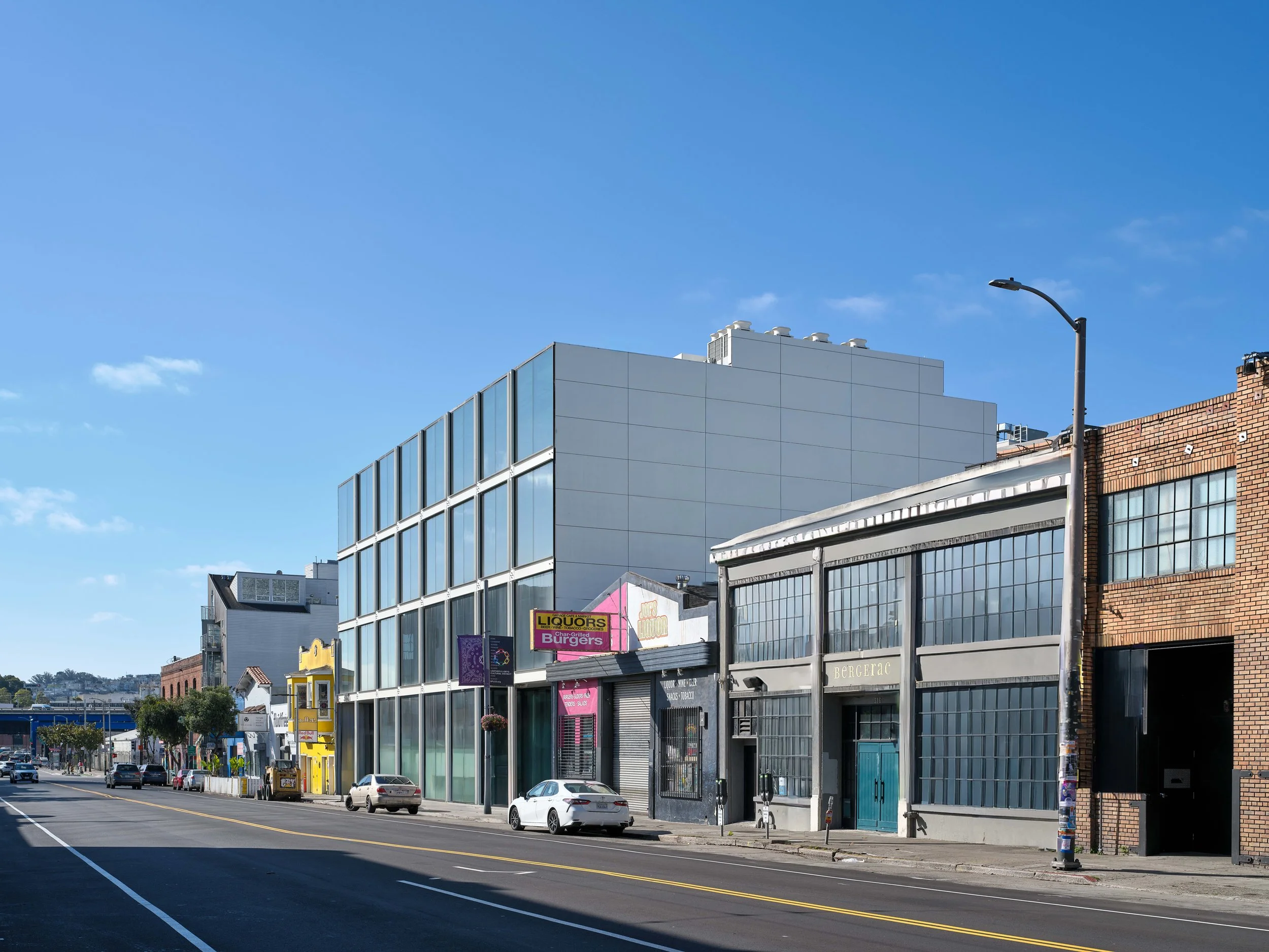 City street with modern glass building, older brick building, parked cars, and streetlamp under a clear blue sky.