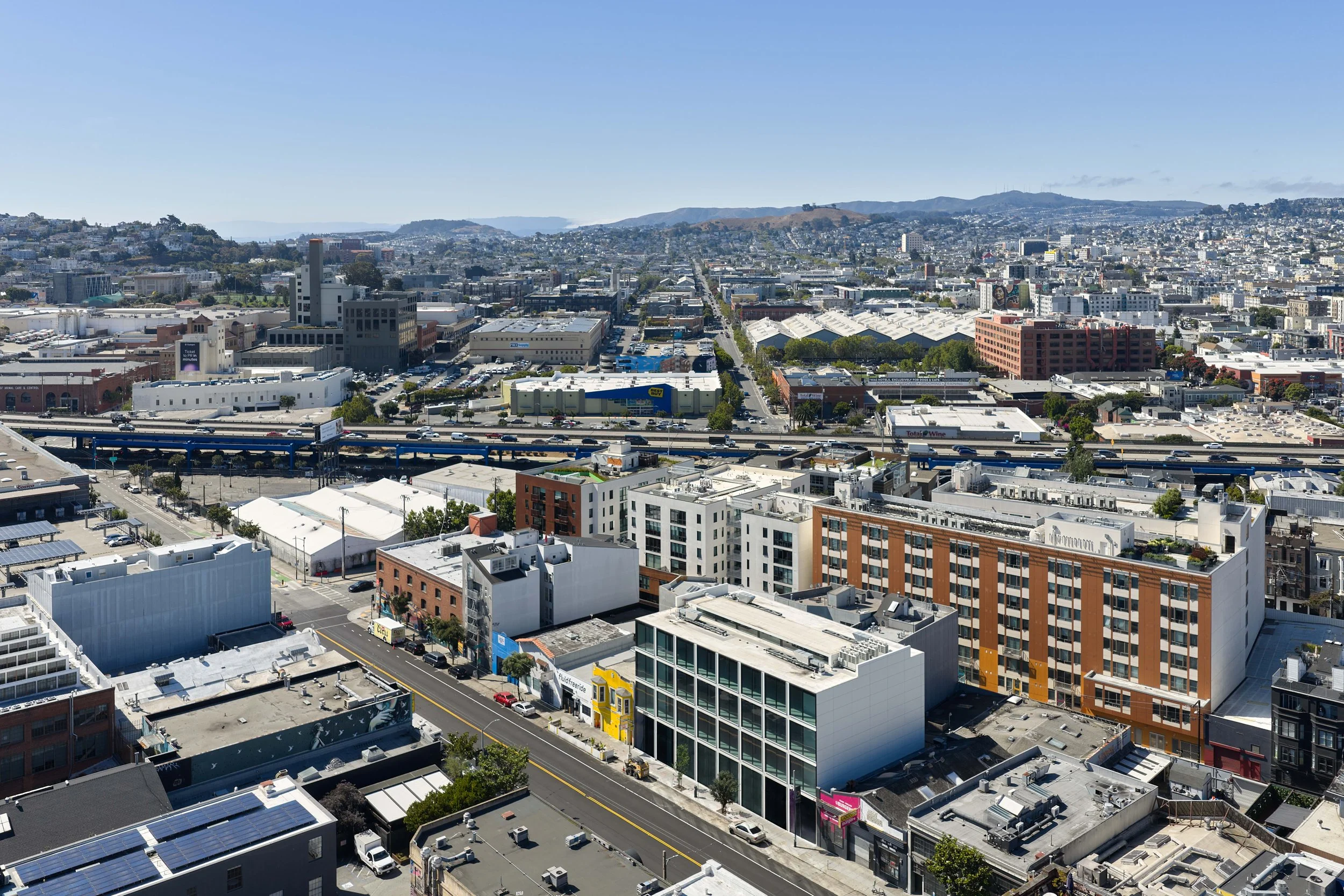 Aerial view of a cityscape with buildings, streets, a highway, and distant hills under a clear blue sky.