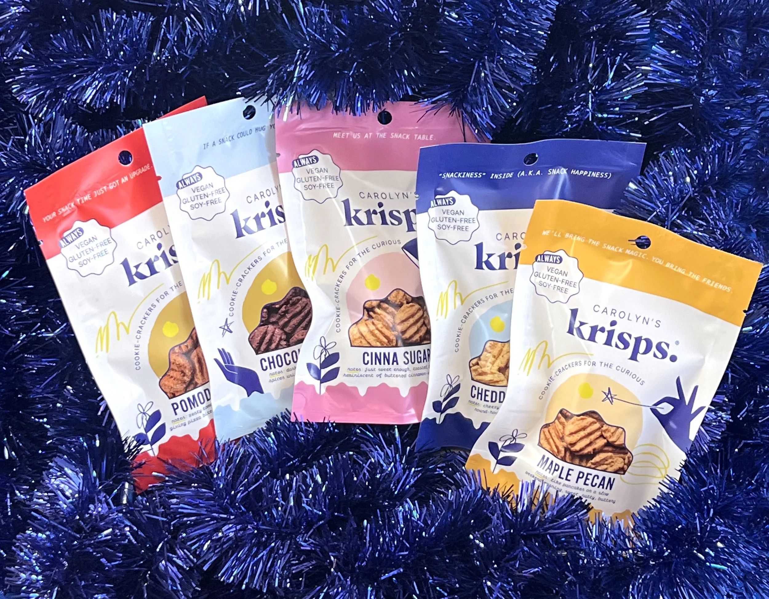 Five bags of Carolynn's Krisps cookie crackers in red, white, pink, blue, and yellow packaging, surrounded by blue tinsel.