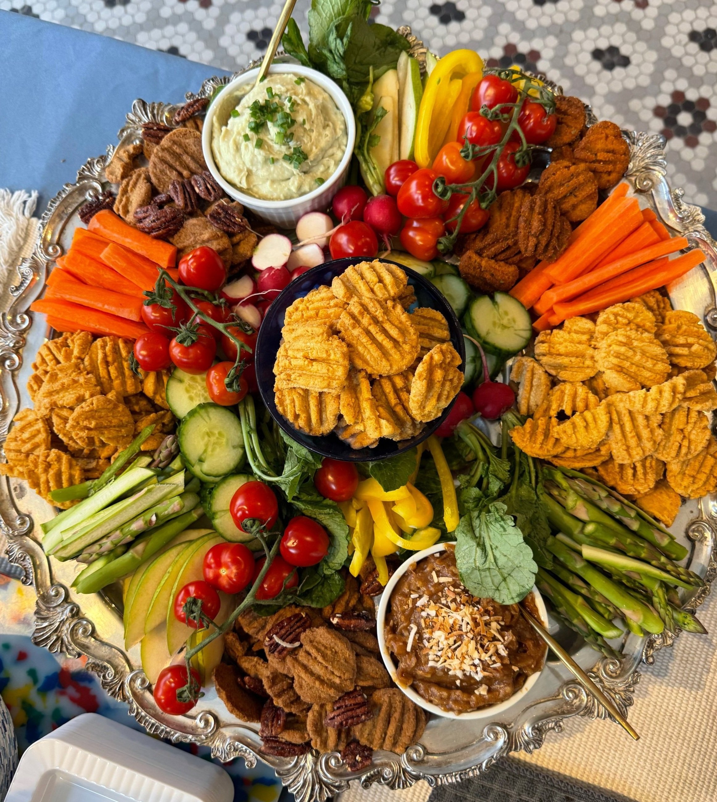 A large silver tray filled with assorted vegetables, crackers, and dips, including cherry tomatoes, radishes, cucumber slices, carrot sticks, pita chips, and bowls of hummus, bean dip, and guacamole.