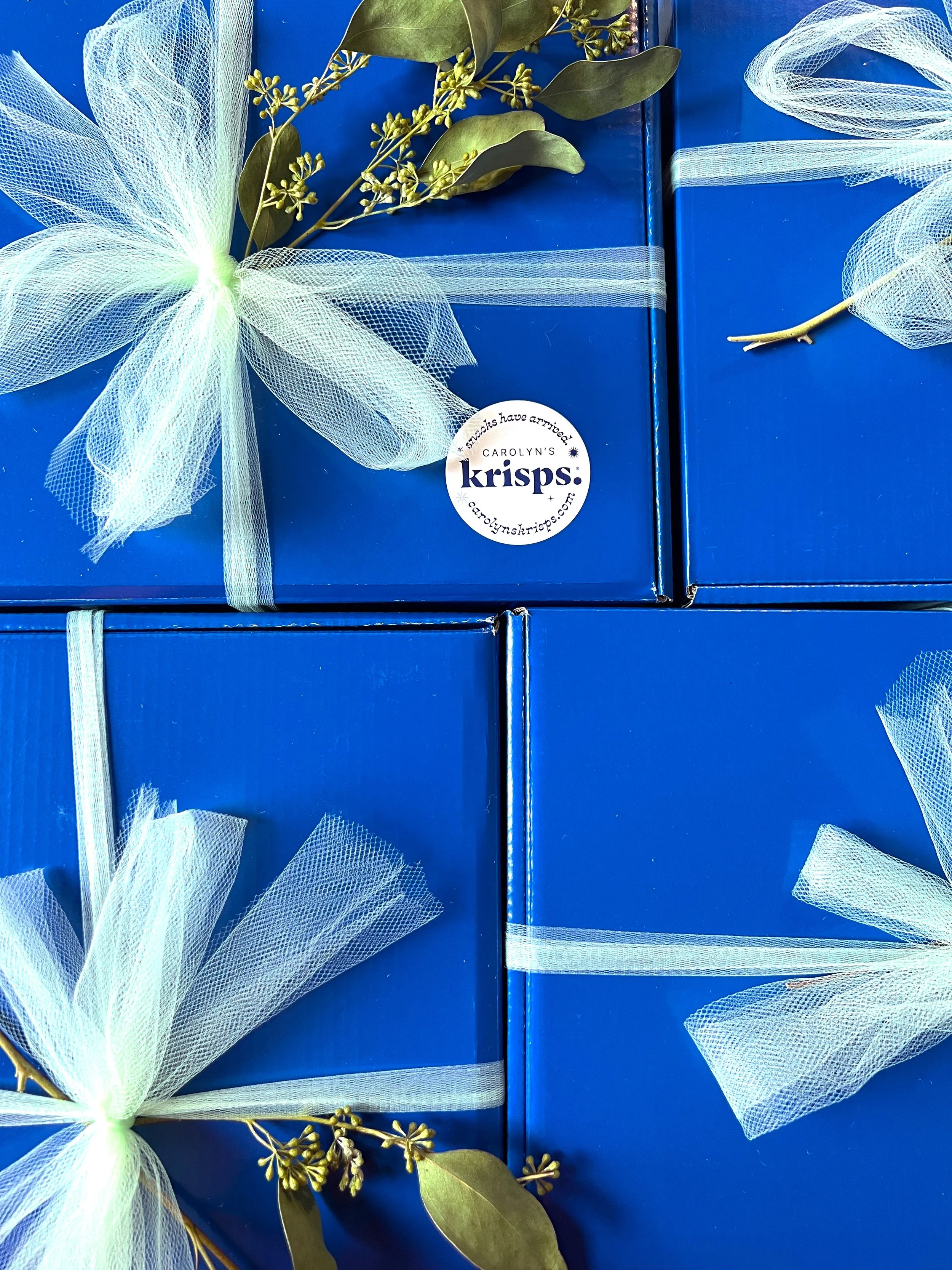 Three blue gift boxes decorated with white mesh ribbons and artificial green leaves with small flowers.