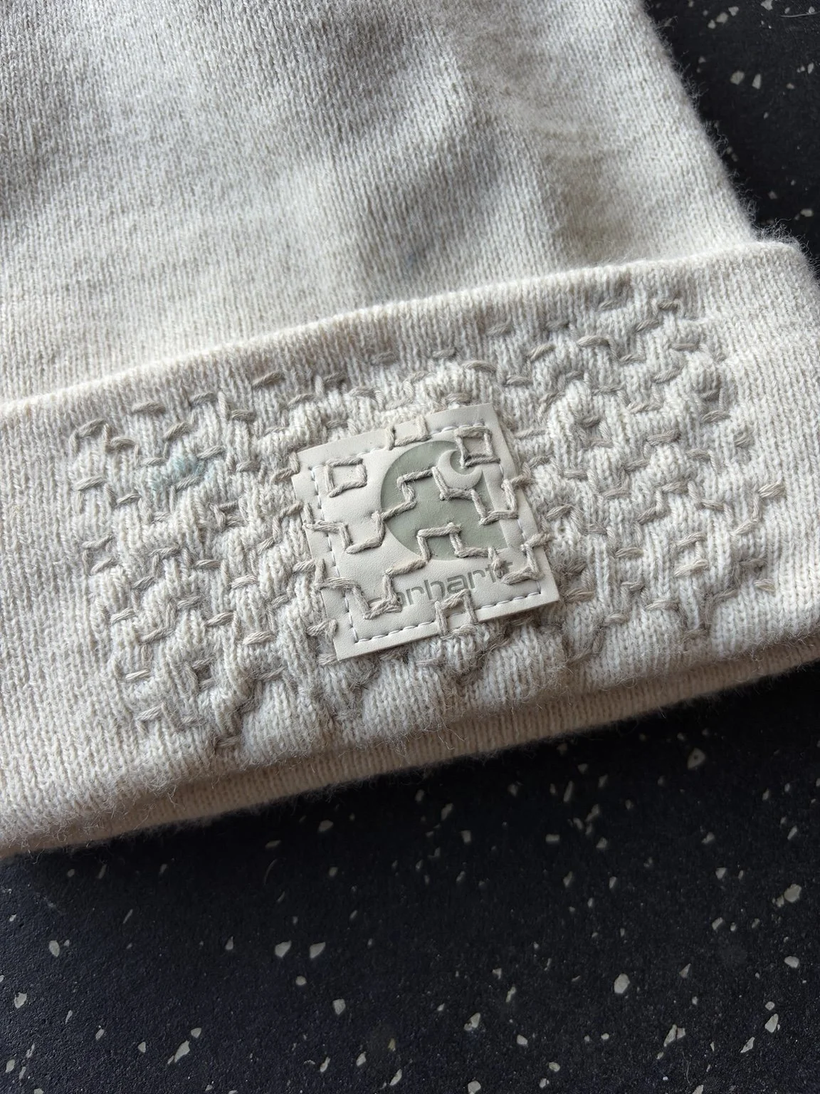 Close-up of tonal sashiko hand stitching crossing the folded cuff and Carhartt patch on Dustin Young’s cream knit beanie.