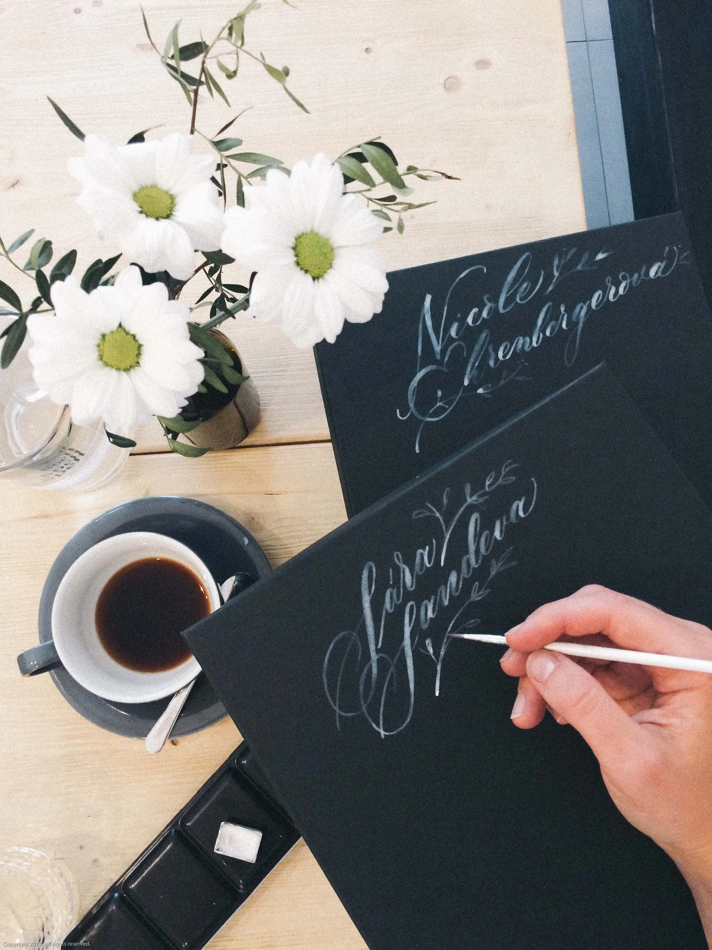 A person writing calligraphy on a black chalkboard, with floral arrangements and a cup of coffee on a light wooden table.