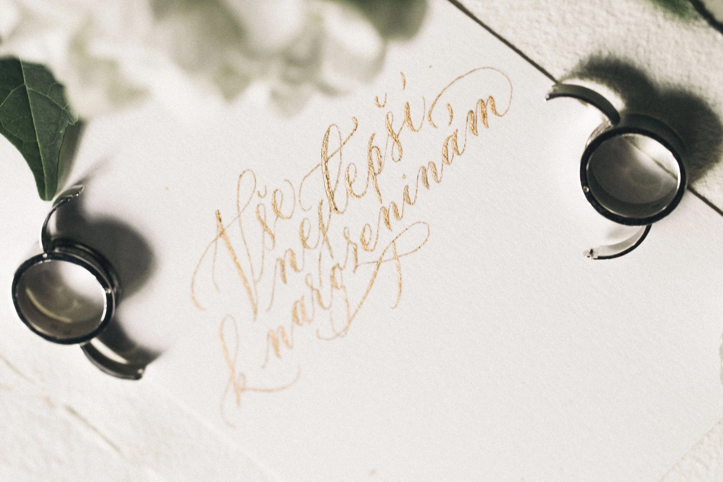 Wedding rings placed on a white paper with calligraphy text that reads 'Hạ sĩ Đặng Thị Phượng and Nguyễn Thị Mai'. Green leaves and white flowers are partially visible in the corners.