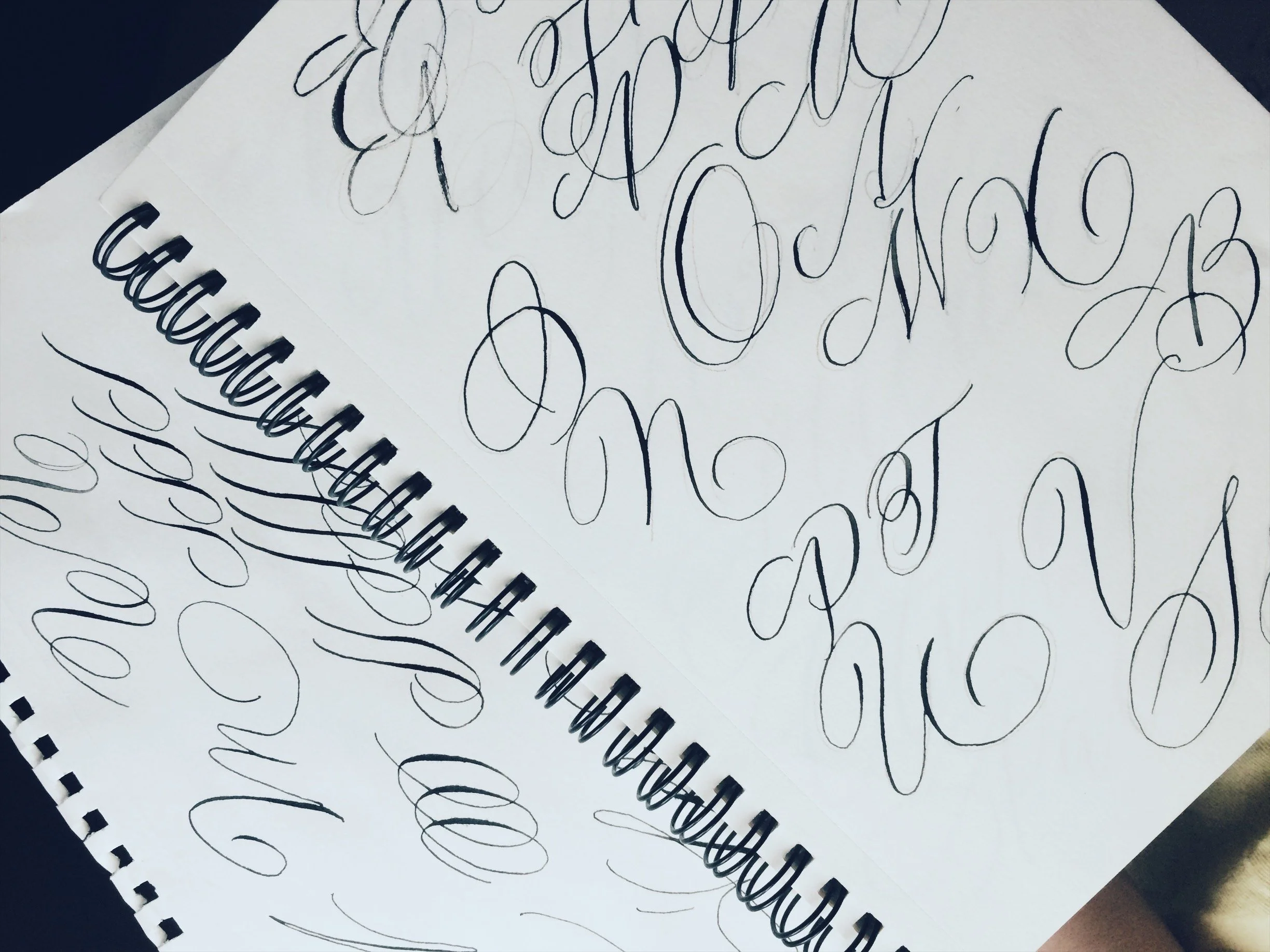 Spiral notebook with black ink cursive calligraphy practice, featuring large swirling letter strokes, and faint practice outlines on white paper.
