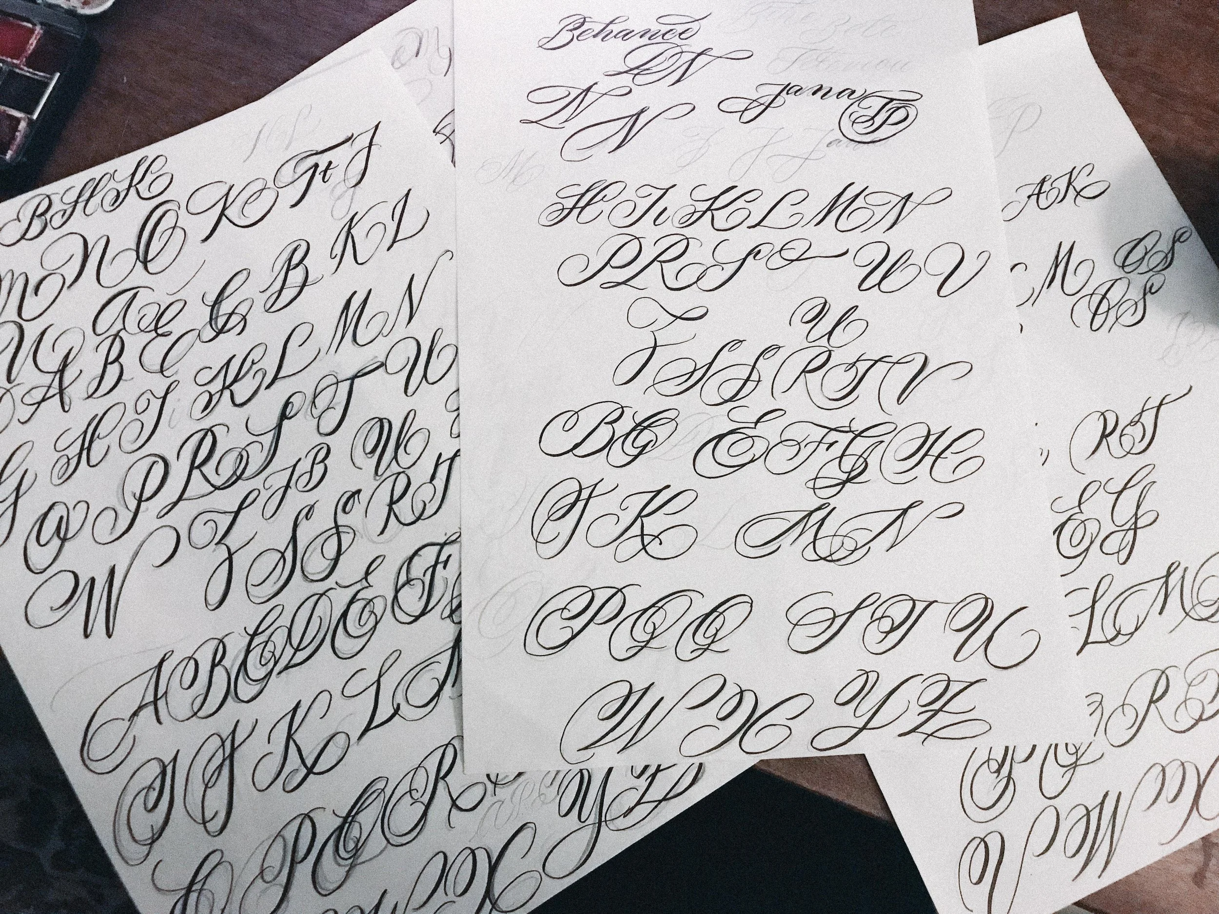 Multiple sheets of white paper with calligraphy alphabet in cursive, arranged on a tabletop.