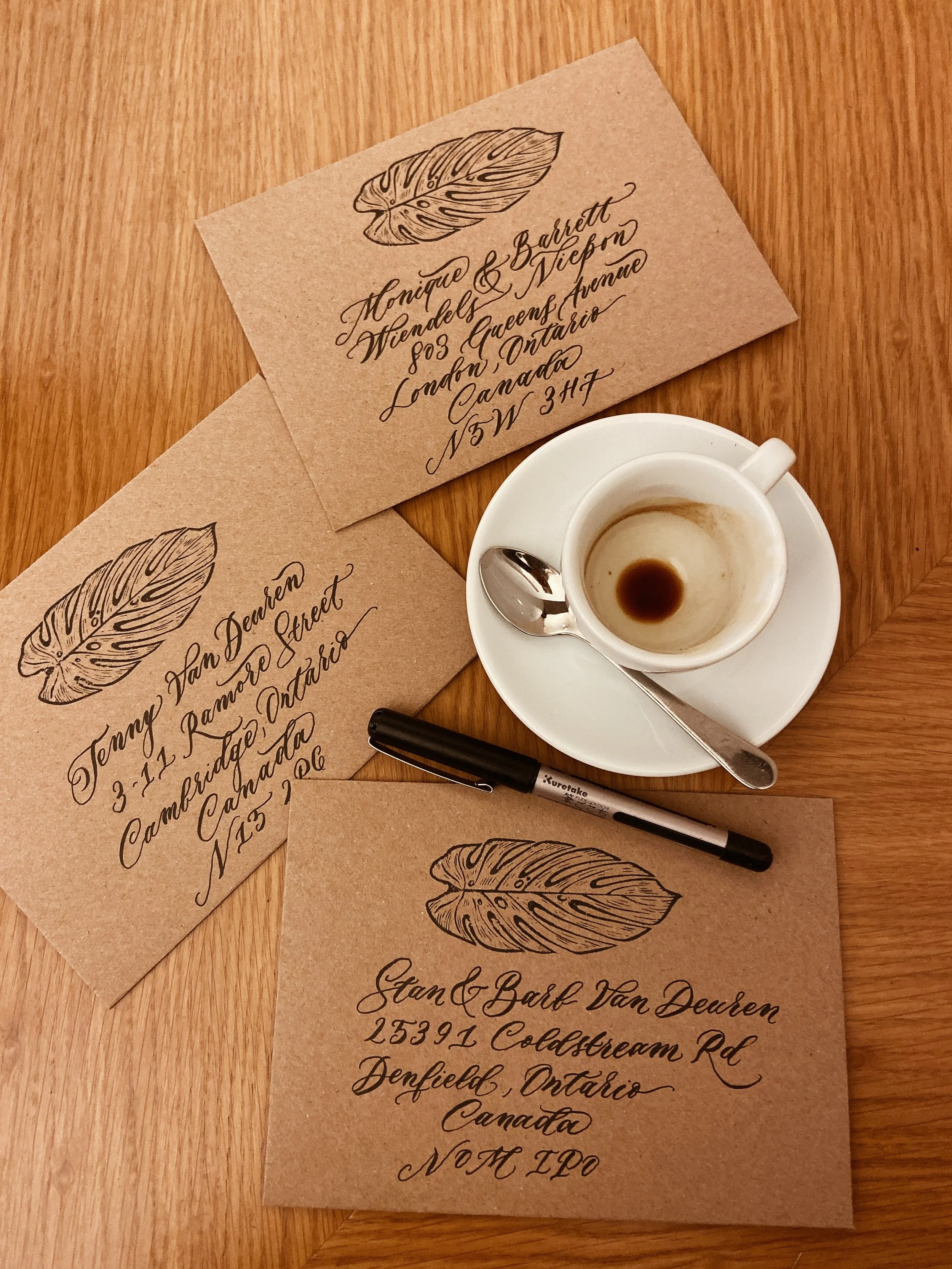 Three envelopes with handwritten calligraphy addresses and a drawing of a leaf on each, placed on a wooden table. One envelope is next to a white coffee cup with a metal spoon, and there is a black marker lying on the table.