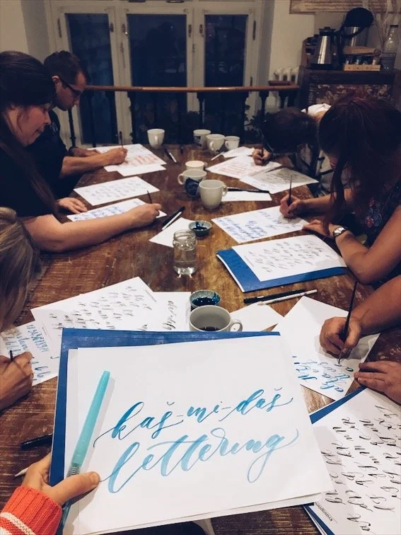 A group of people gathered around a wooden table practicing calligraphy with papers, pens, and cups of coffee or tea.