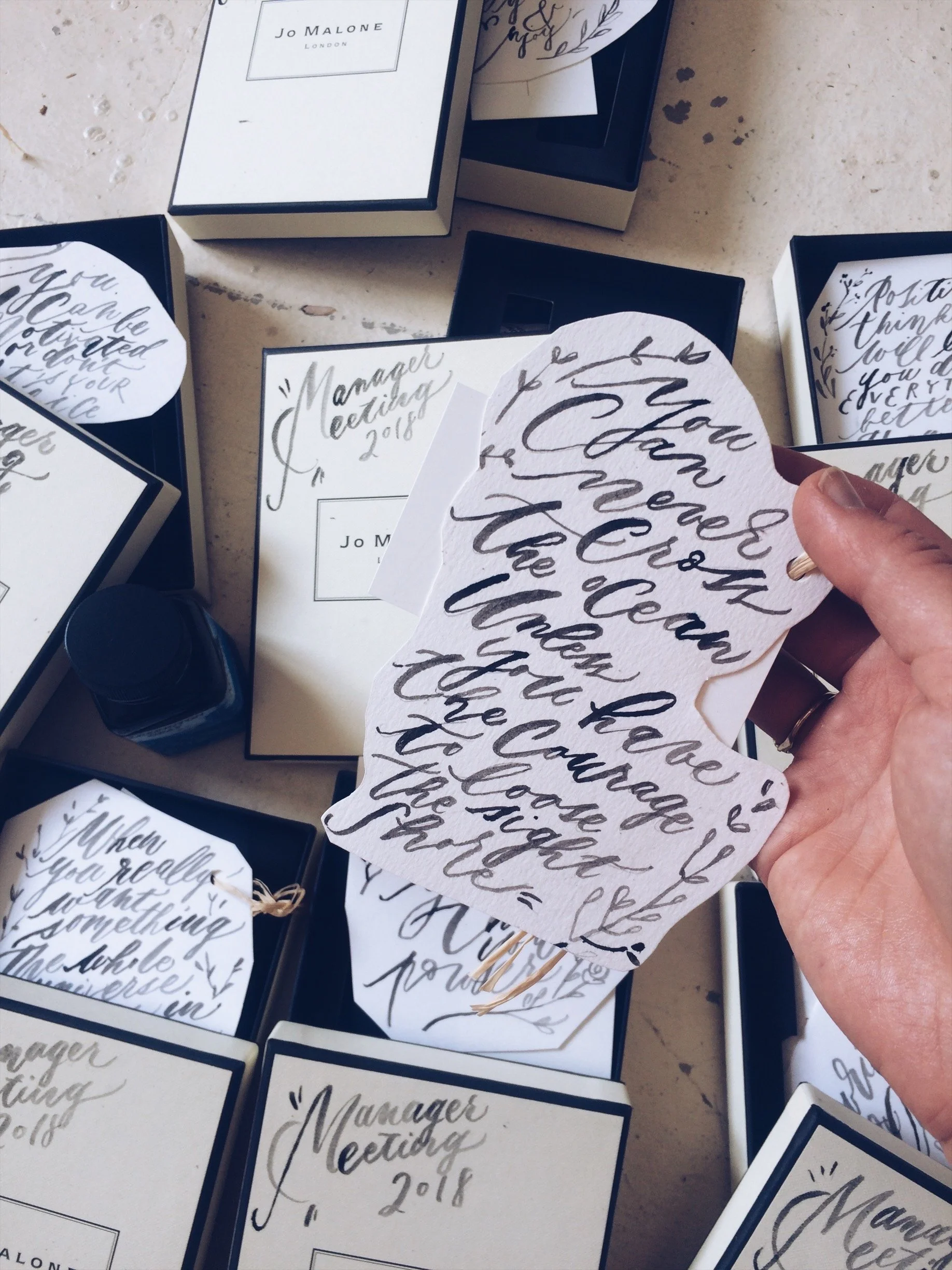 Person holding a decorative paper with calligraphy words, among various boxes of similar papers and a bottle of ink on a table.