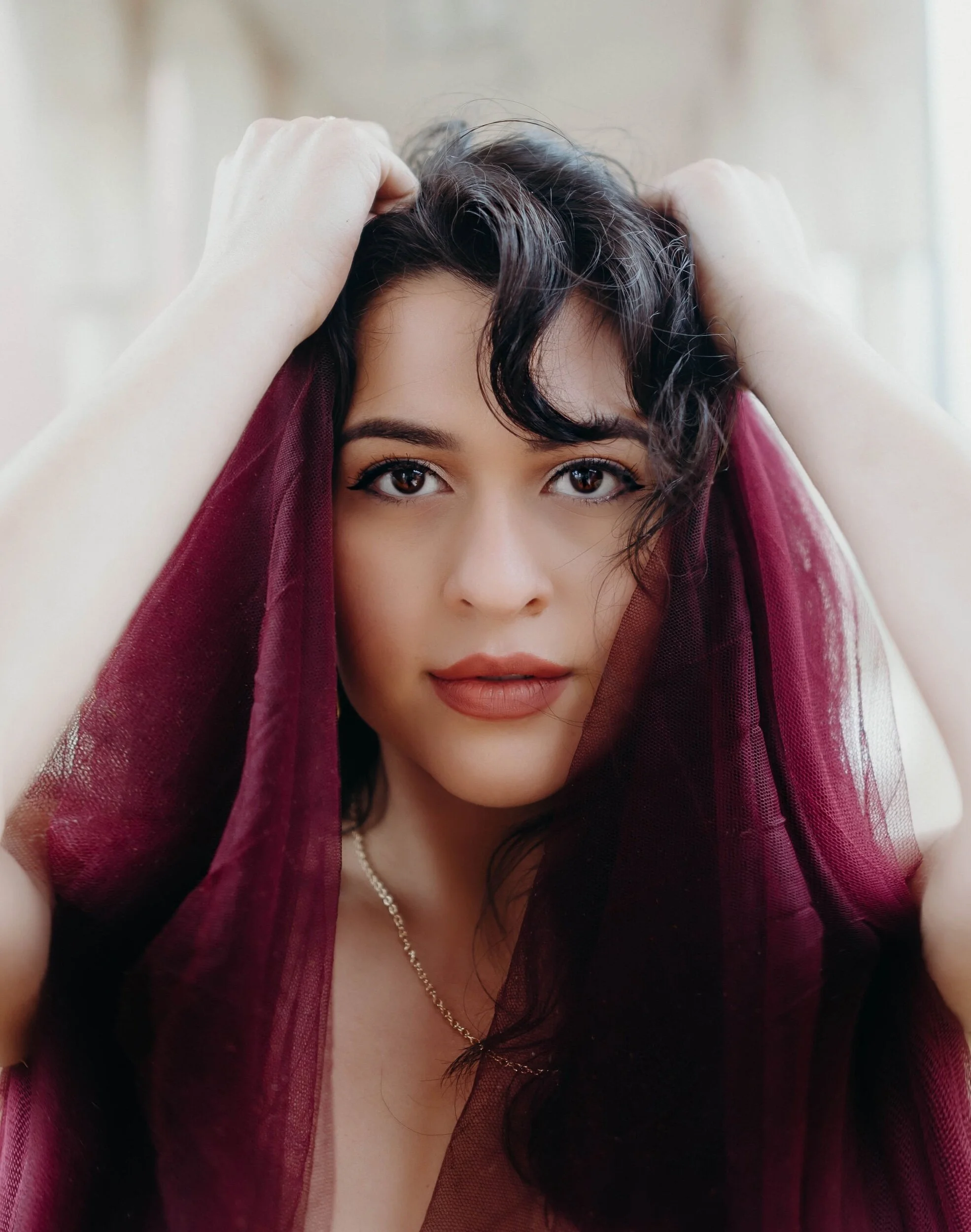 Close-up of a woman with dark wavy hair, holding her hair with both hands, partially covered with maroon sheer fabric, looking directly at the camera.