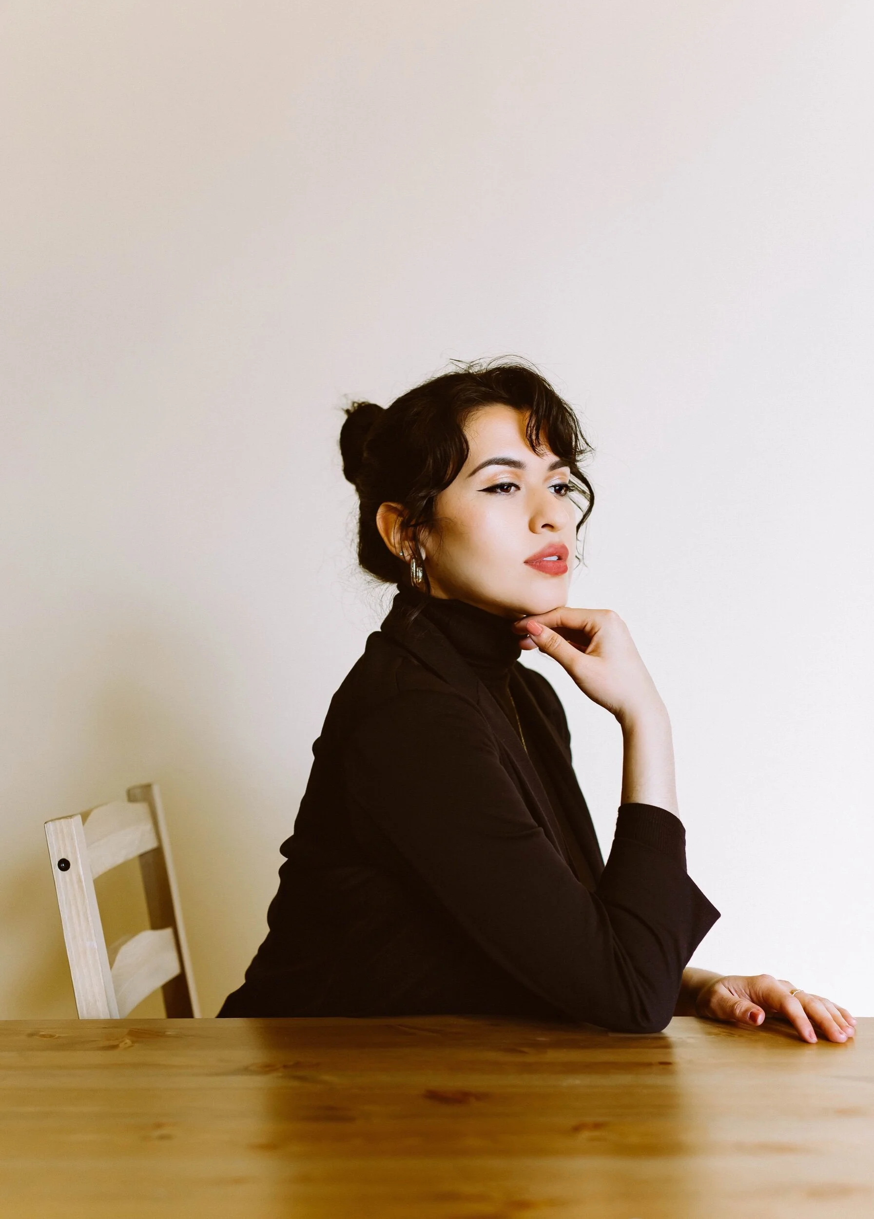 A woman with dark hair styled in a bun, wearing a black turtleneck and earrings, sitting at a wooden table against a plain white wall.