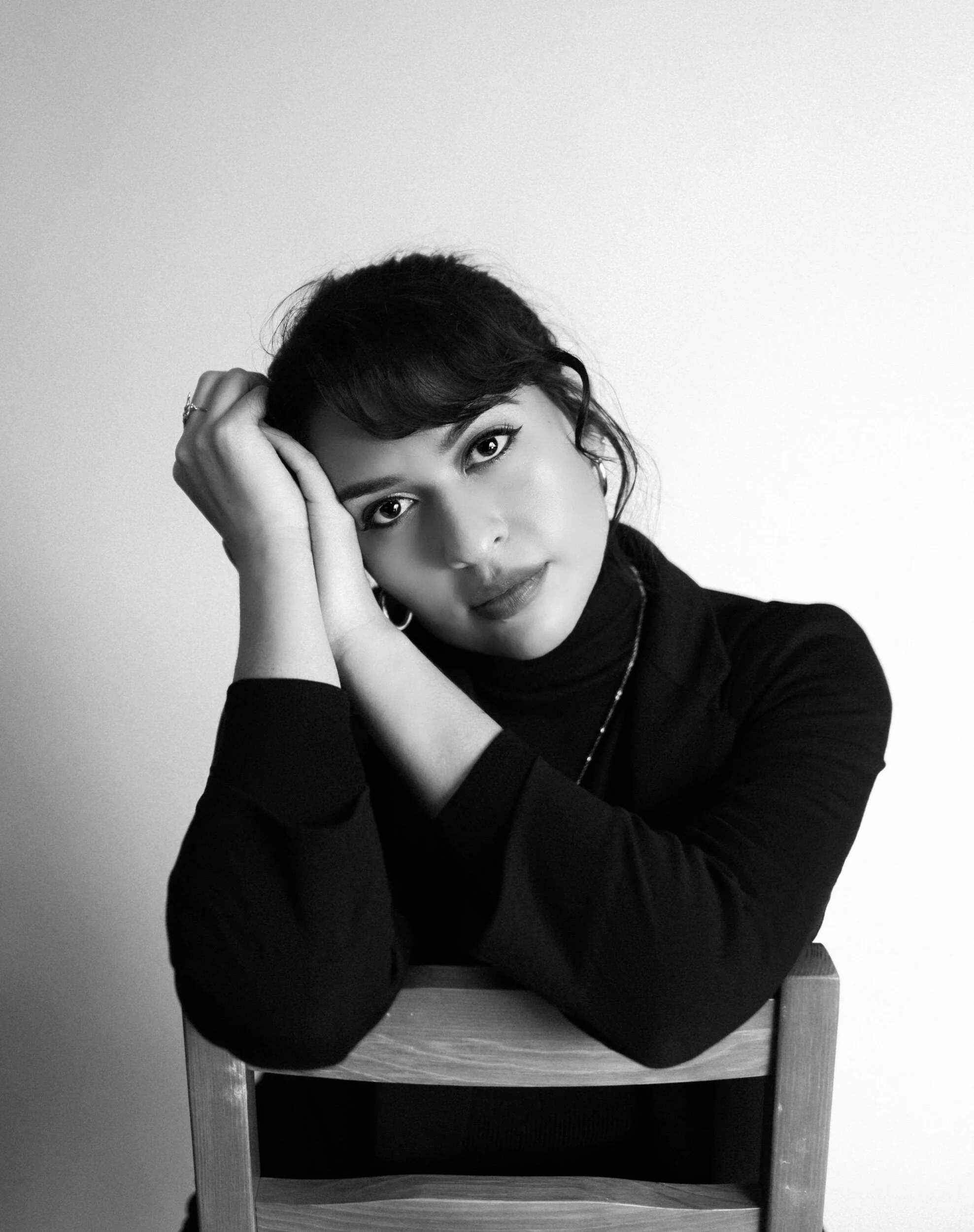 Black and white portrait of a young woman with dark hair and light eyes, sitting with her arms resting on the back of a wooden chair, looking directly at the camera.