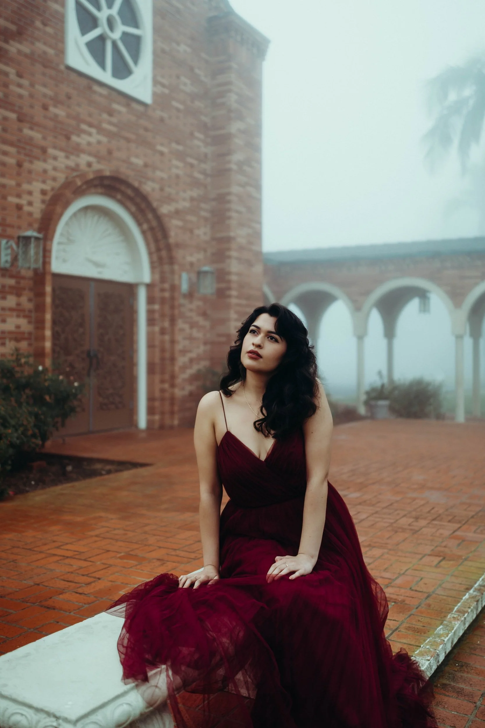 A woman in a deep red gown sitting on a stone ledge outside a brick building with a church-like architecture, foggy atmosphere, and palm trees in the background.