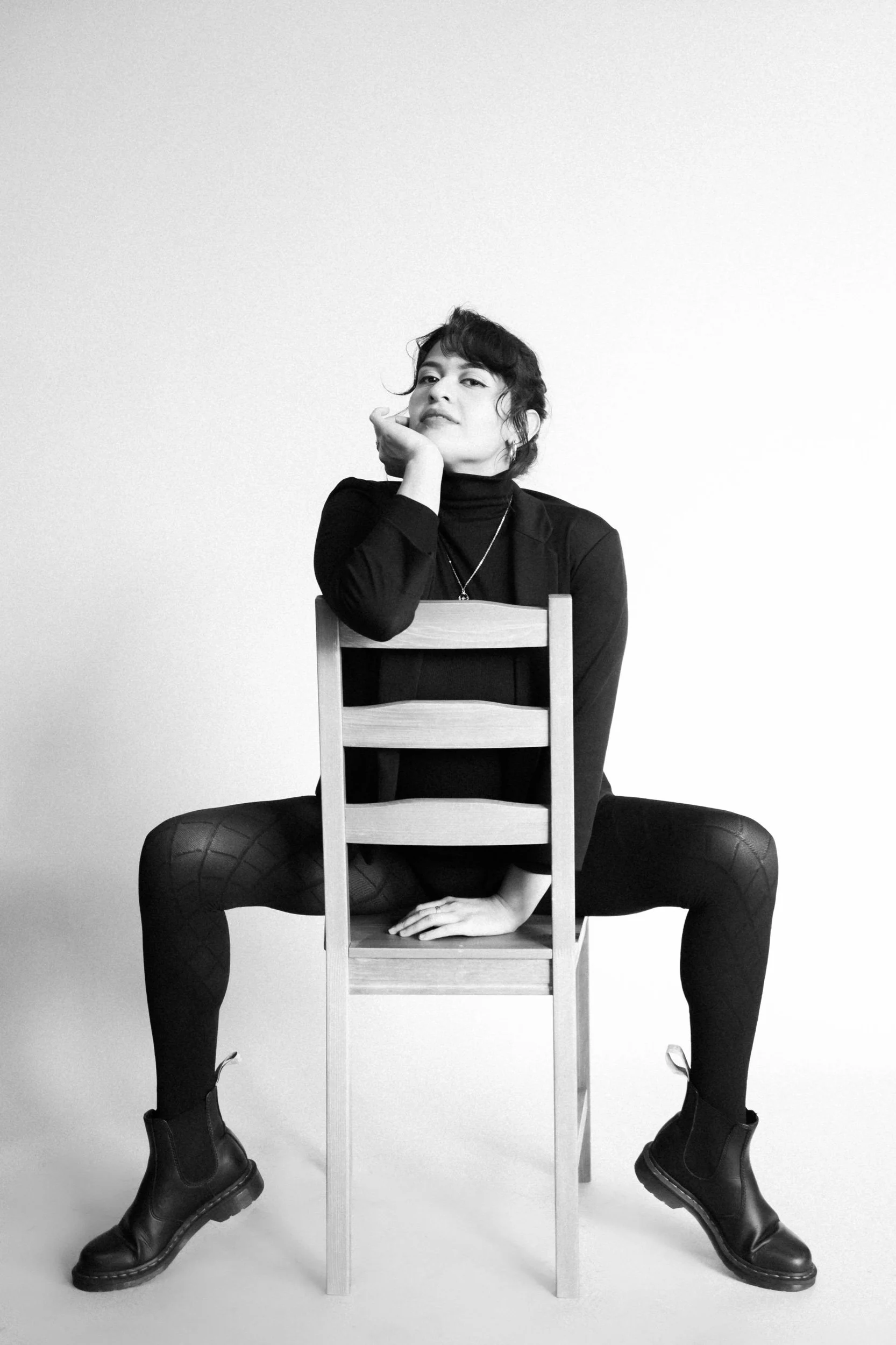 Black and white photo of a woman sitting on a chair with her legs apart, wearing a black turtleneck, blazer, tights with a diamond pattern, and combat boots, resting her chin on her hand with a confident expression