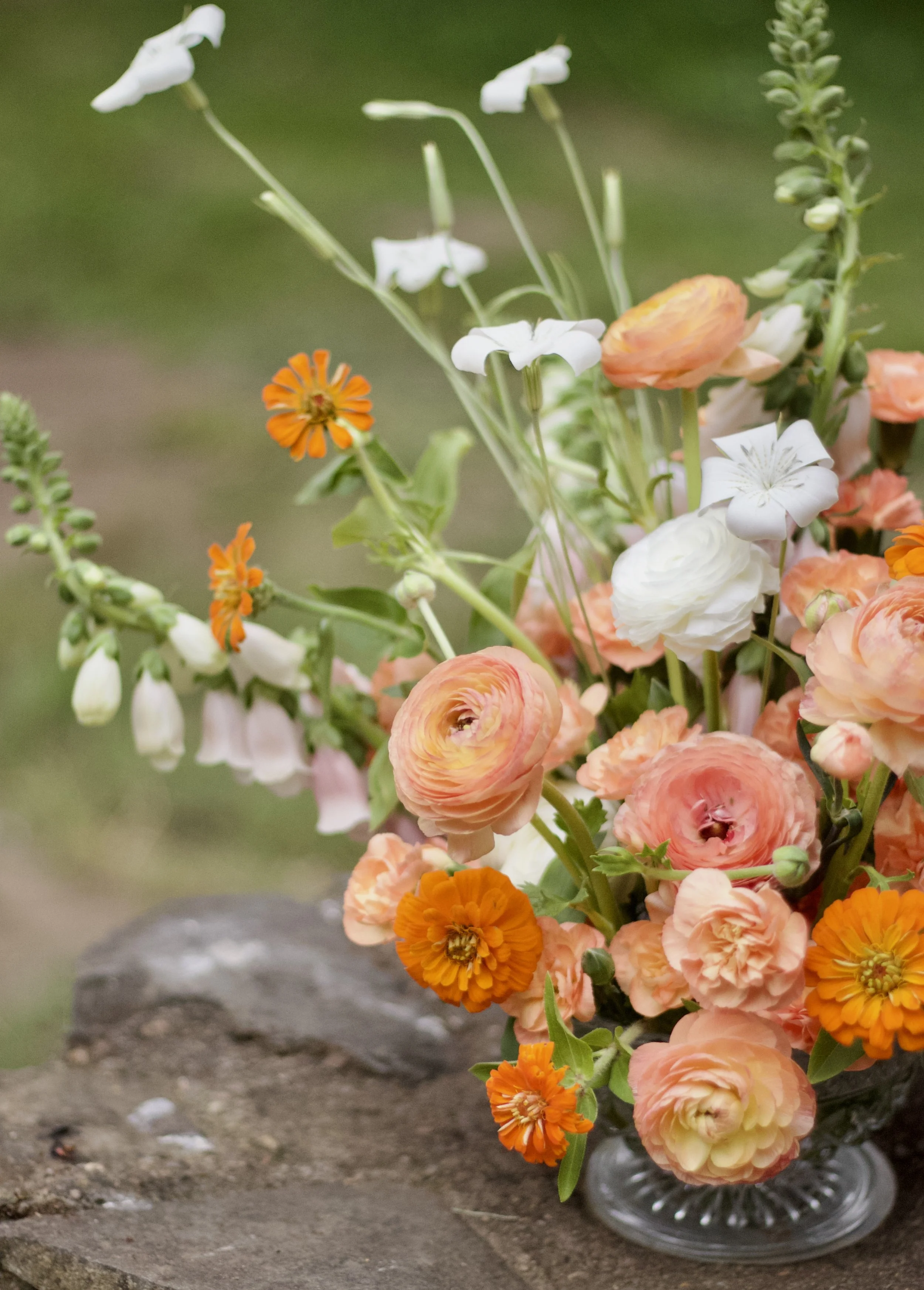 A close-up of a mixed flower bouquet in a glass vase, featuring orange, peach, and white flowers, against a blurred outdoor background.