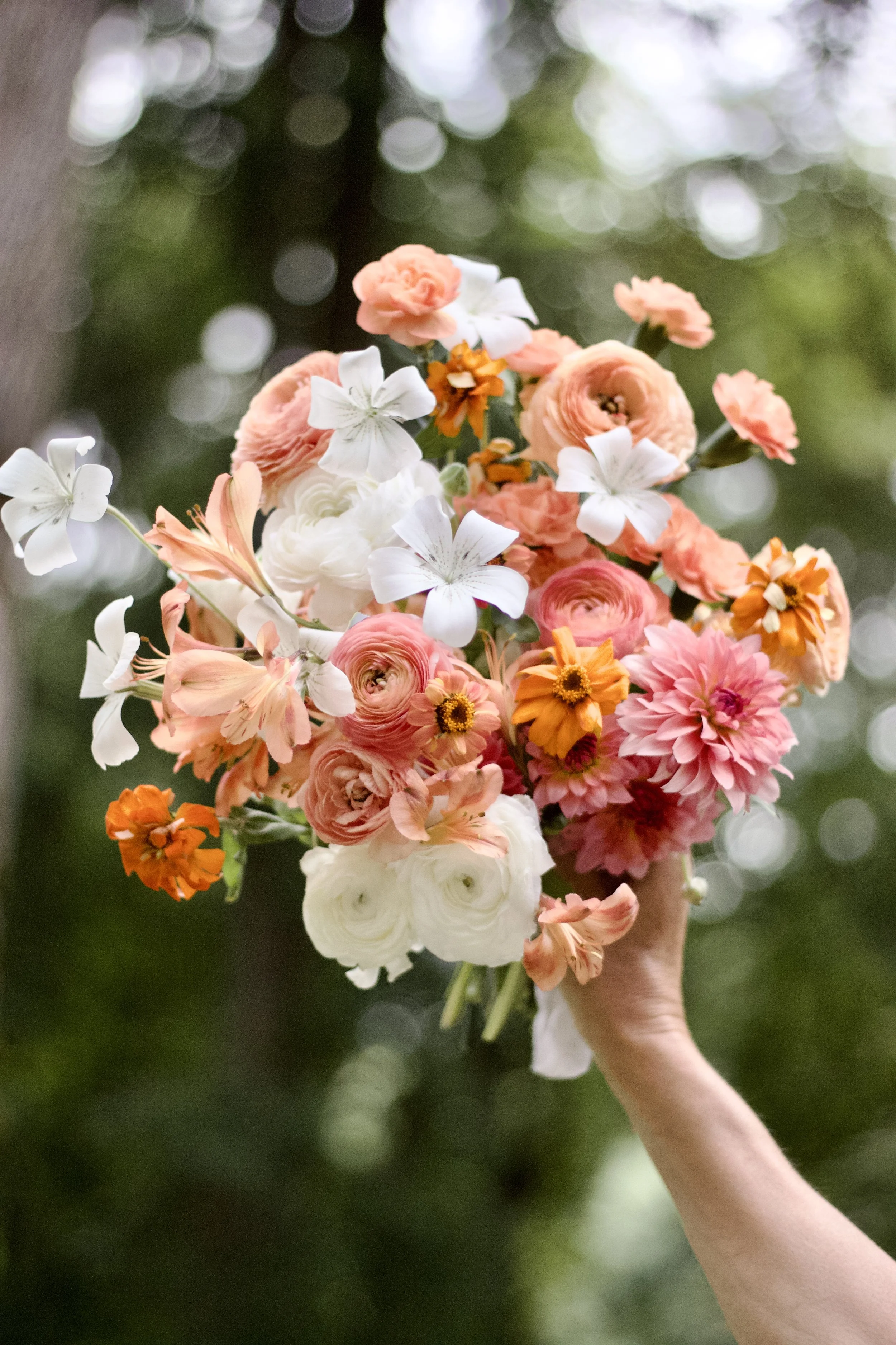 A hand holding a bouquet of various pastel-colored flowers, including white, peach, and pink blooms, against a blurred outdoor green background.