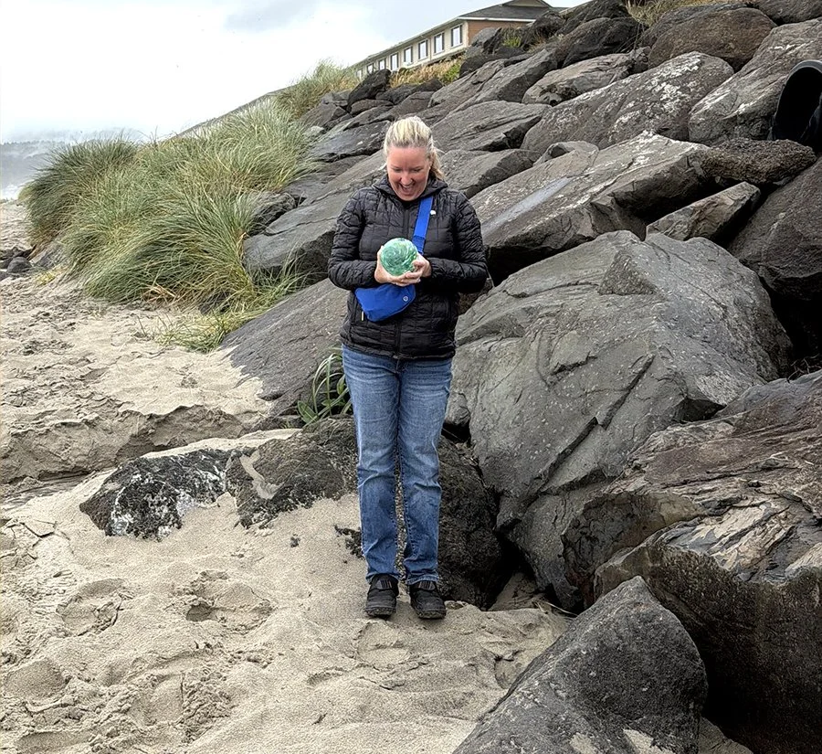 woman in black jacket and blue jeans holding a glass float, standing on the beach next to large rocks