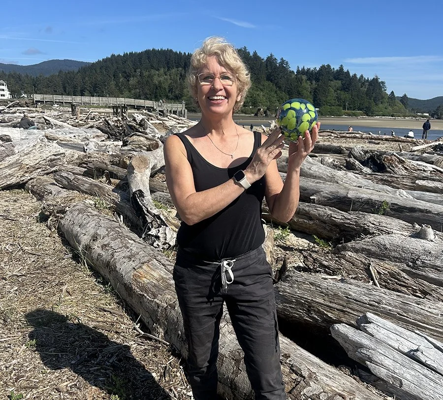 women holding a glass float standing next to driftwood on a beach