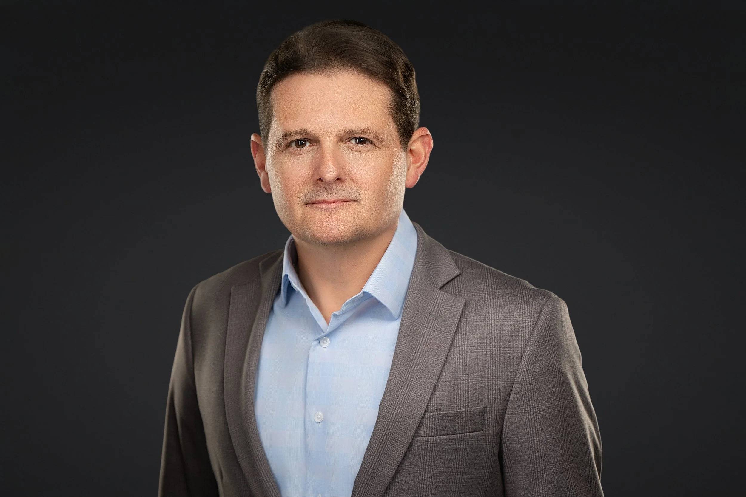 Headshot of a middle-aged man with brown hair, wearing a gray suit and blue shirt, against a dark background.