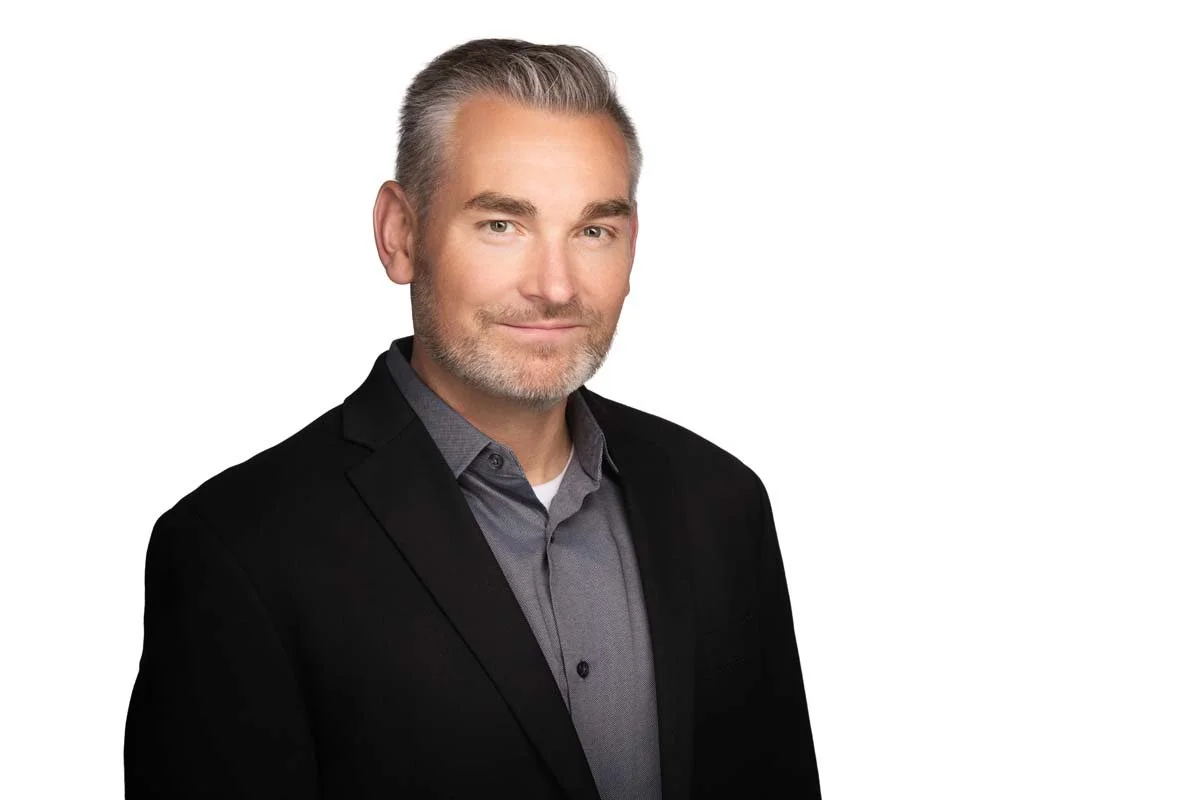 A middle-aged man with gray hair and a beard, wearing a black blazer over a gray shirt, smiling softly at the camera.