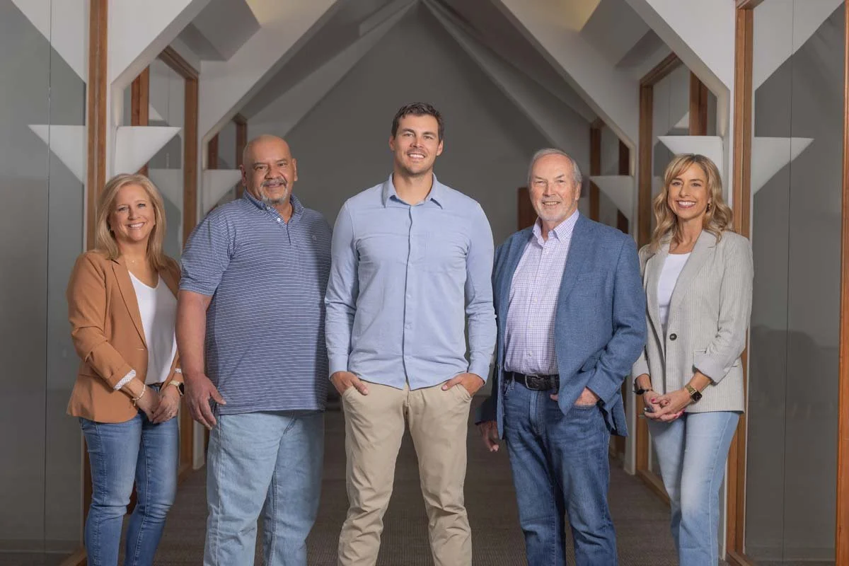 Group of five professionally dressed people standing in hallway with an A-frame ceiling and wooden accents, smiling at camera.