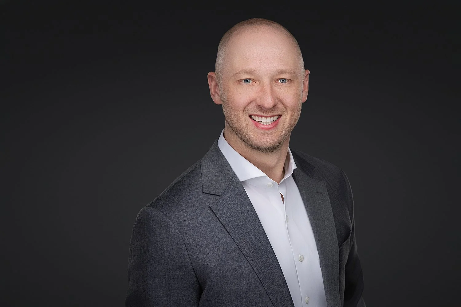 Portrait of a smiling man with a shaved head wearing a gray suit and white shirt against a dark background.