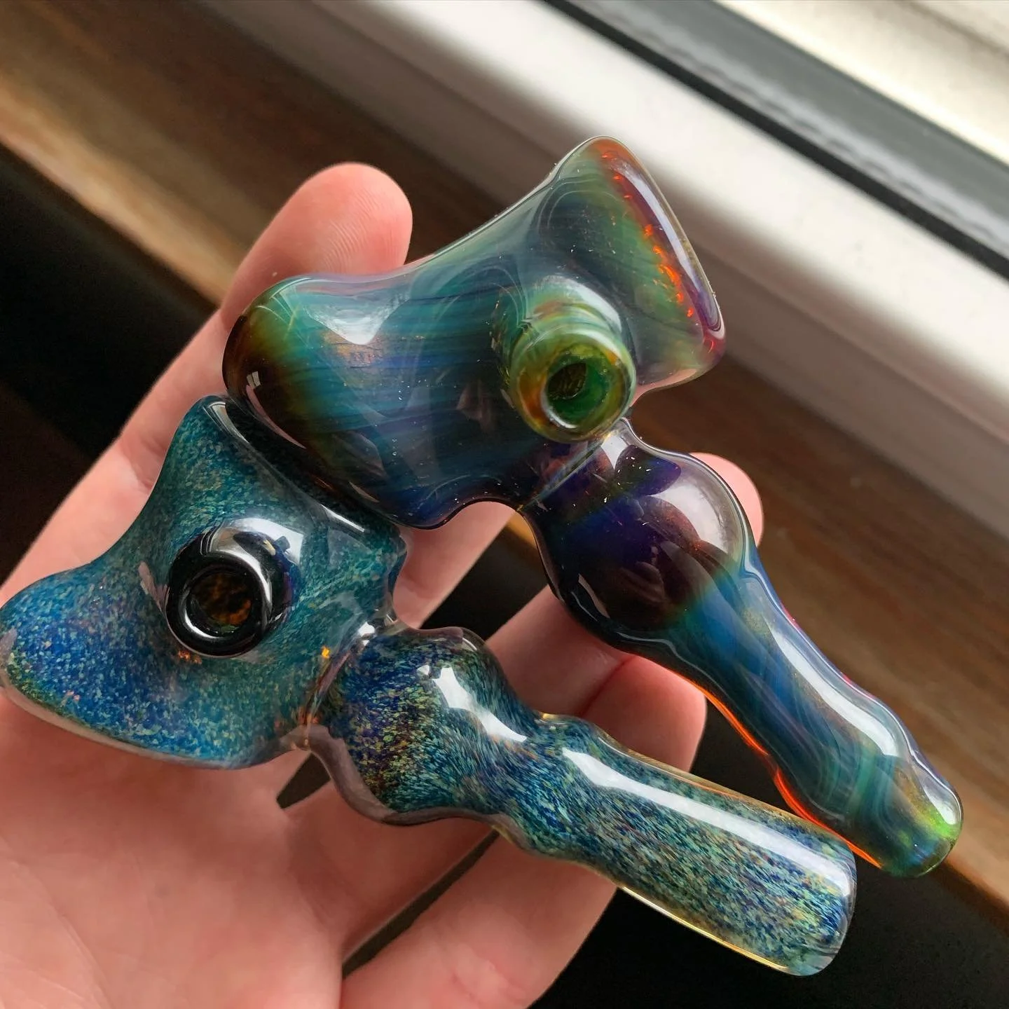 A person holding a colorful, abstract glass pipe with swirls of green, blue, purple, and hints of orange, near a window.