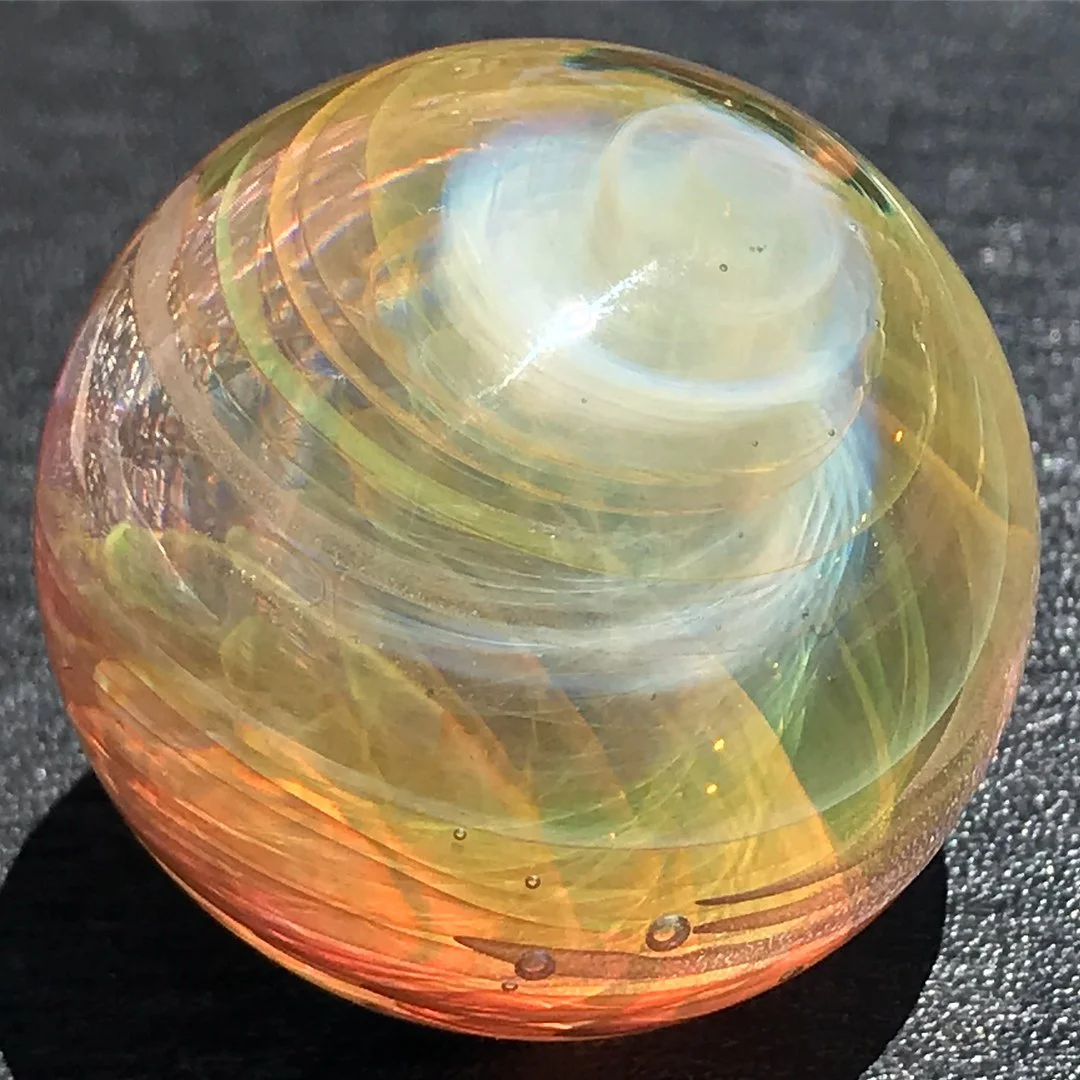 Colorful glass marble with swirling patterns of yellow, white, and hints of pink and green, resting on a black surface.