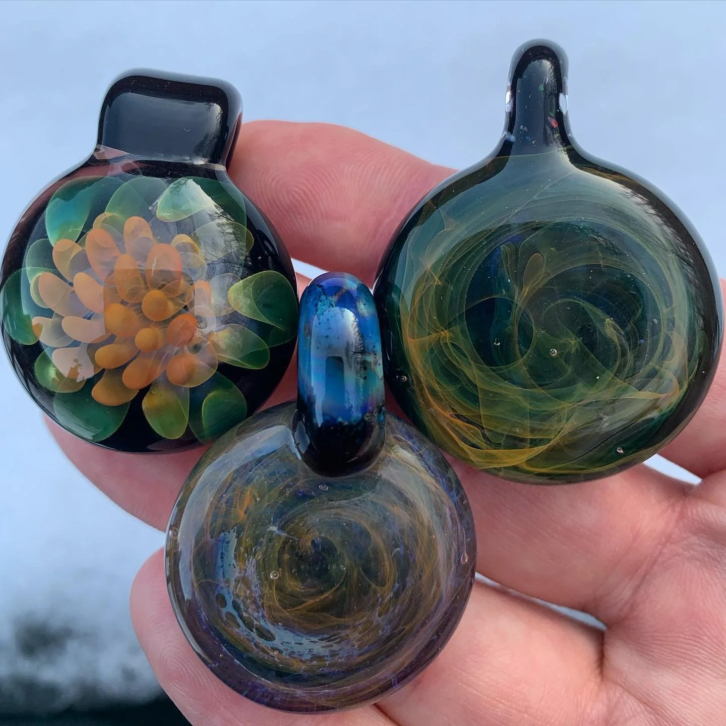Close-up of three colorful glass pendants with floral and abstract designs, being held by a person outdoors.